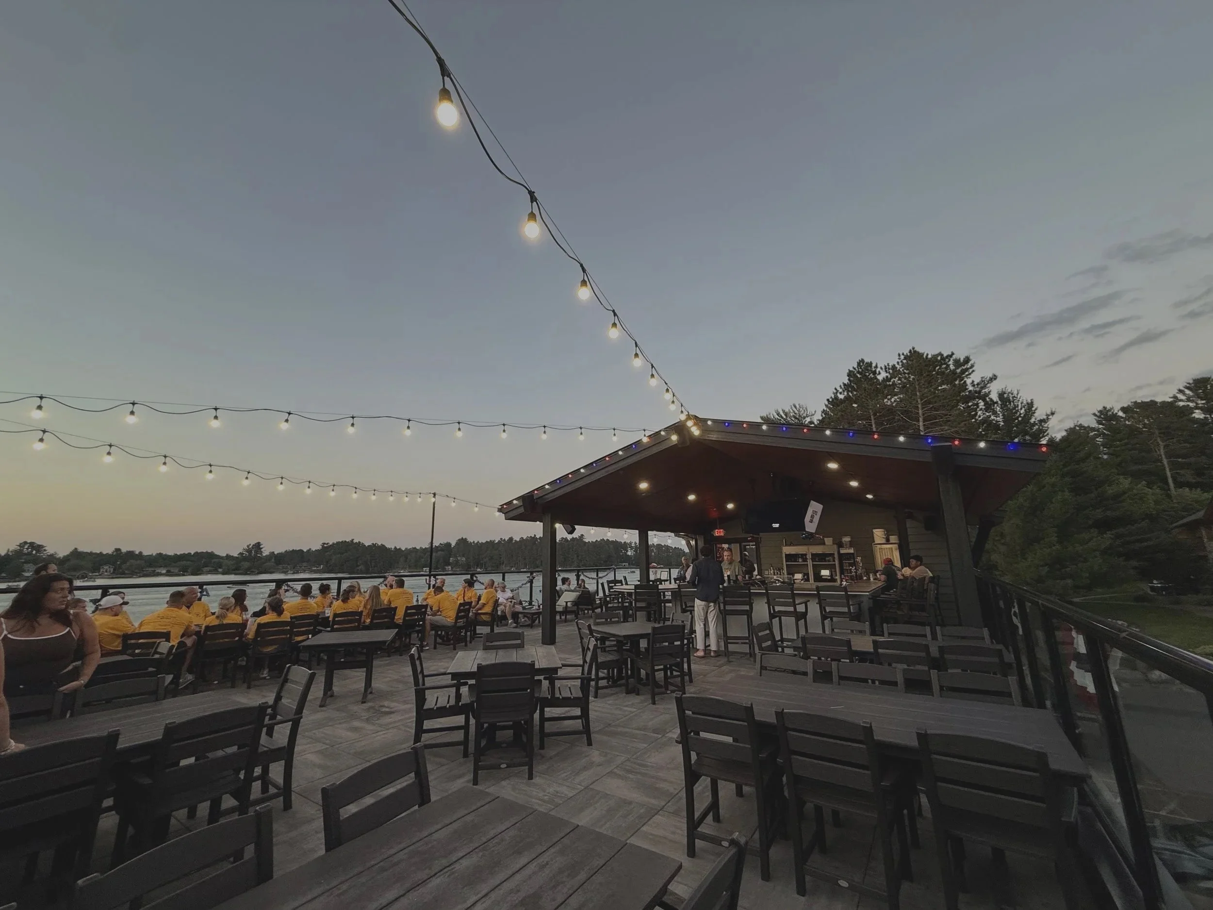 An outdoor patio area with string lights, some people sitting and standing, a building with a roof decorated with small lights, and a body of water in the background during dusk.