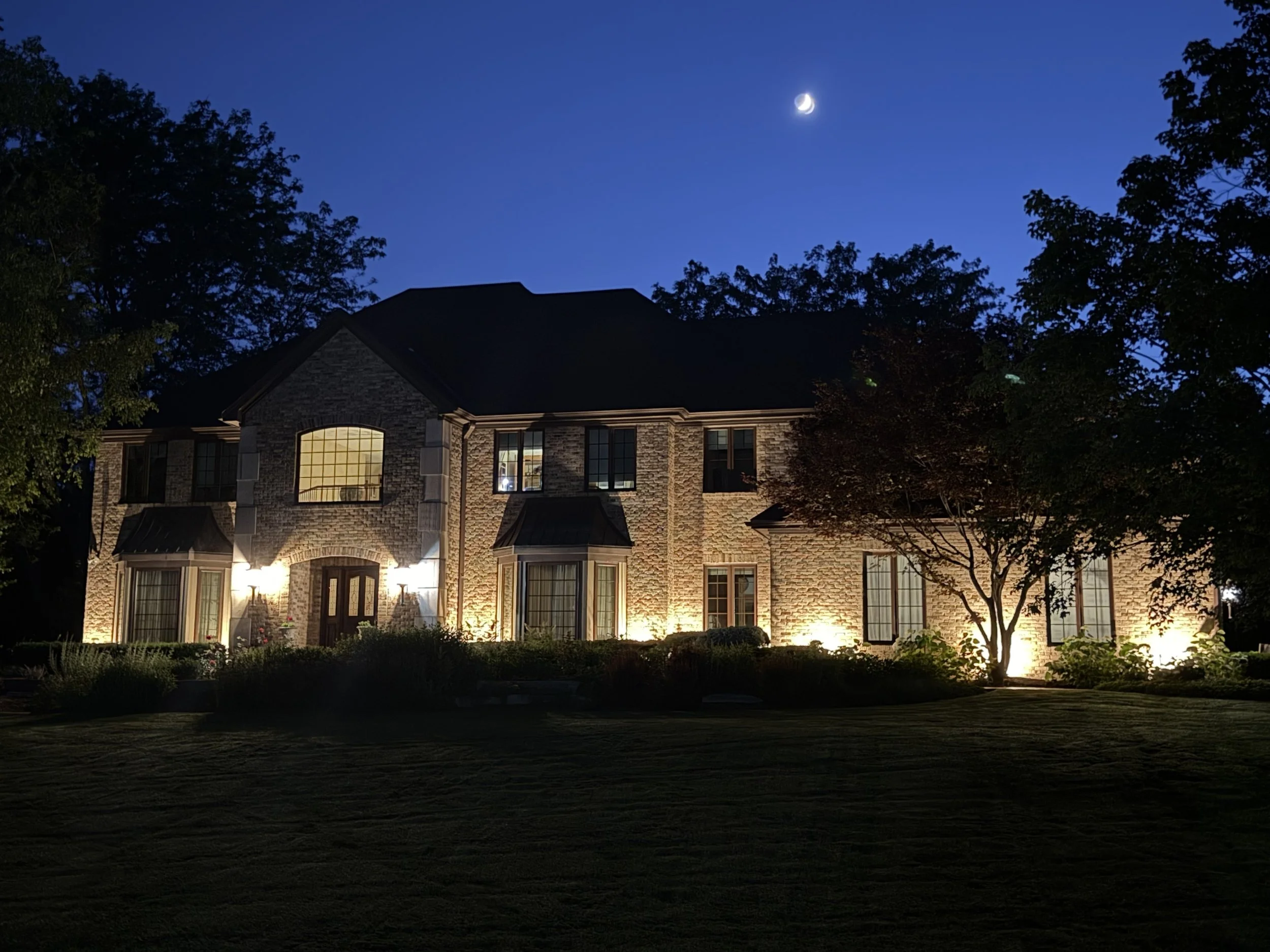 A two-story brick house with lit windows at night, surrounded by trees, under a dark blue sky with a crescent moon.
