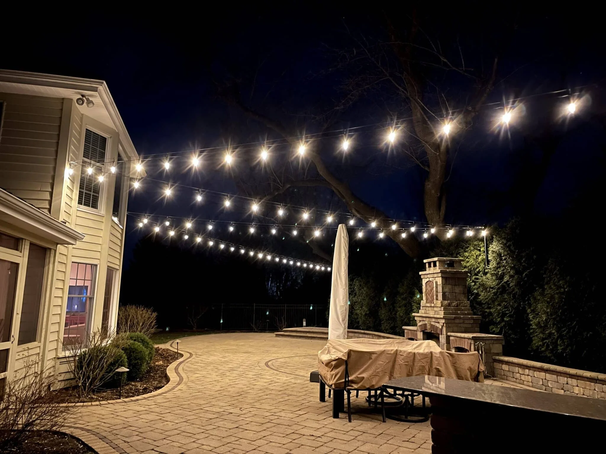 Nighttime view of a backyard patio illuminated by string lights hanging above. The patio has a brick surface, a covered outdoor furniture, and a brick fireplace or grill. A large tree with bare branches is visible behind the lights.