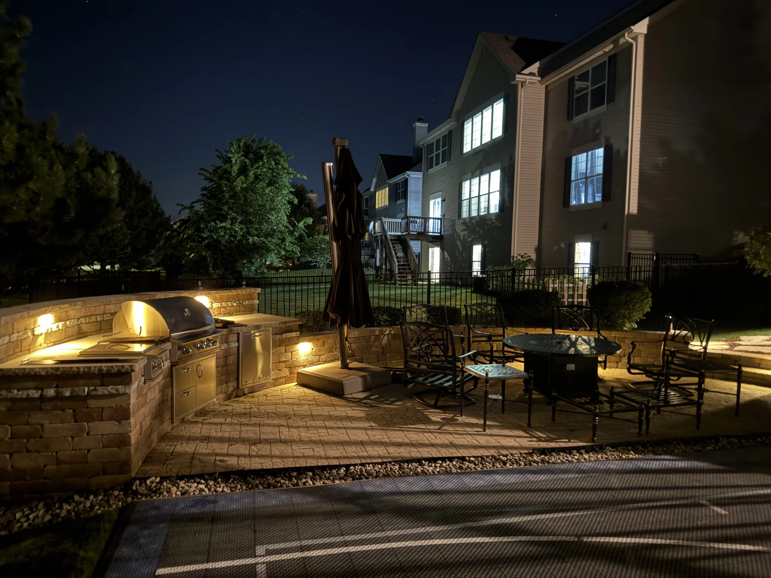 Nighttime backyard patio with lit outdoor grill, patio furniture, fenced yard, trees, large house with lit windows, and a clear starry sky.