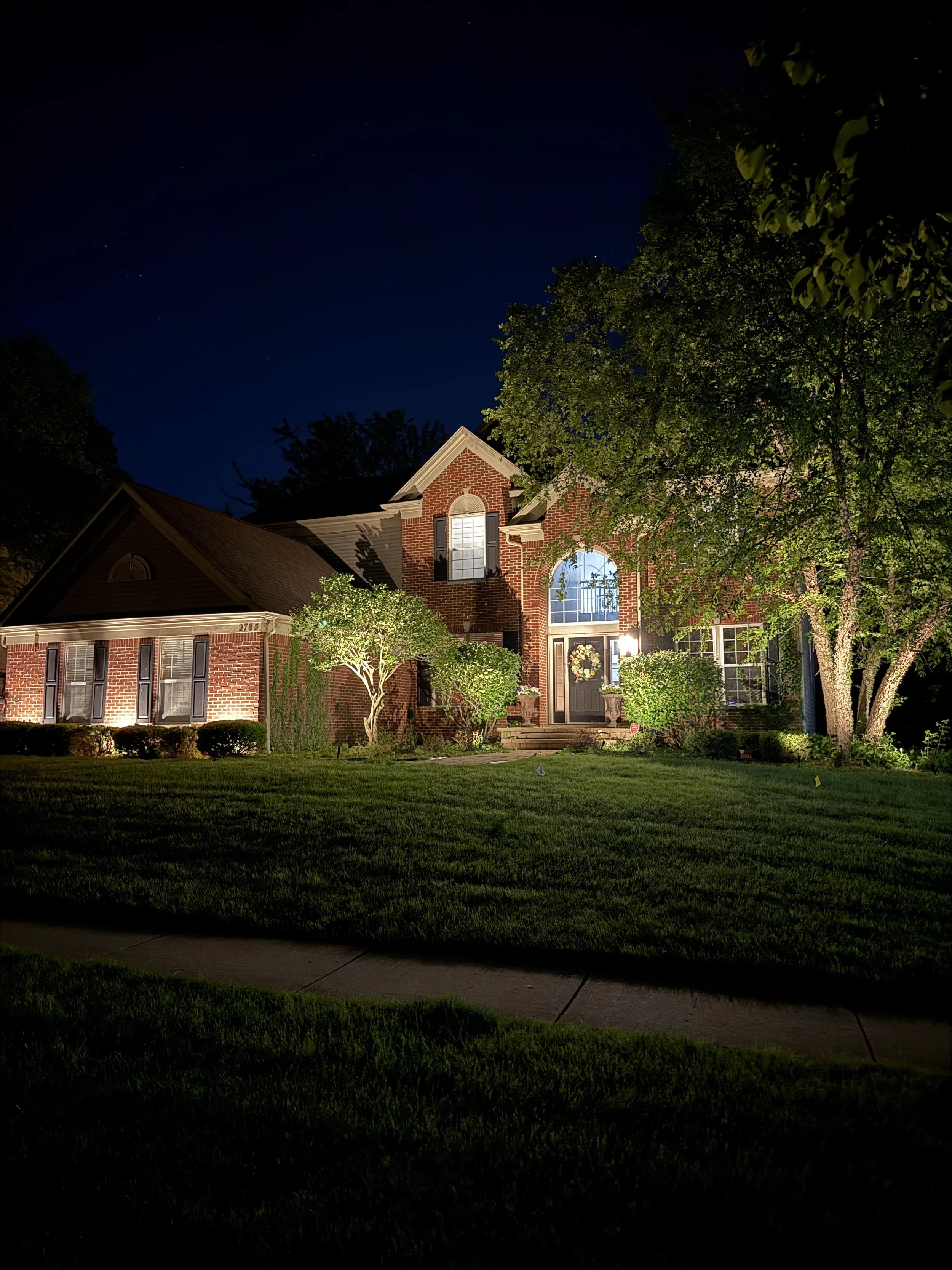 A brick house with lit windows at night, surrounded by green trees and a well-maintained lawn.