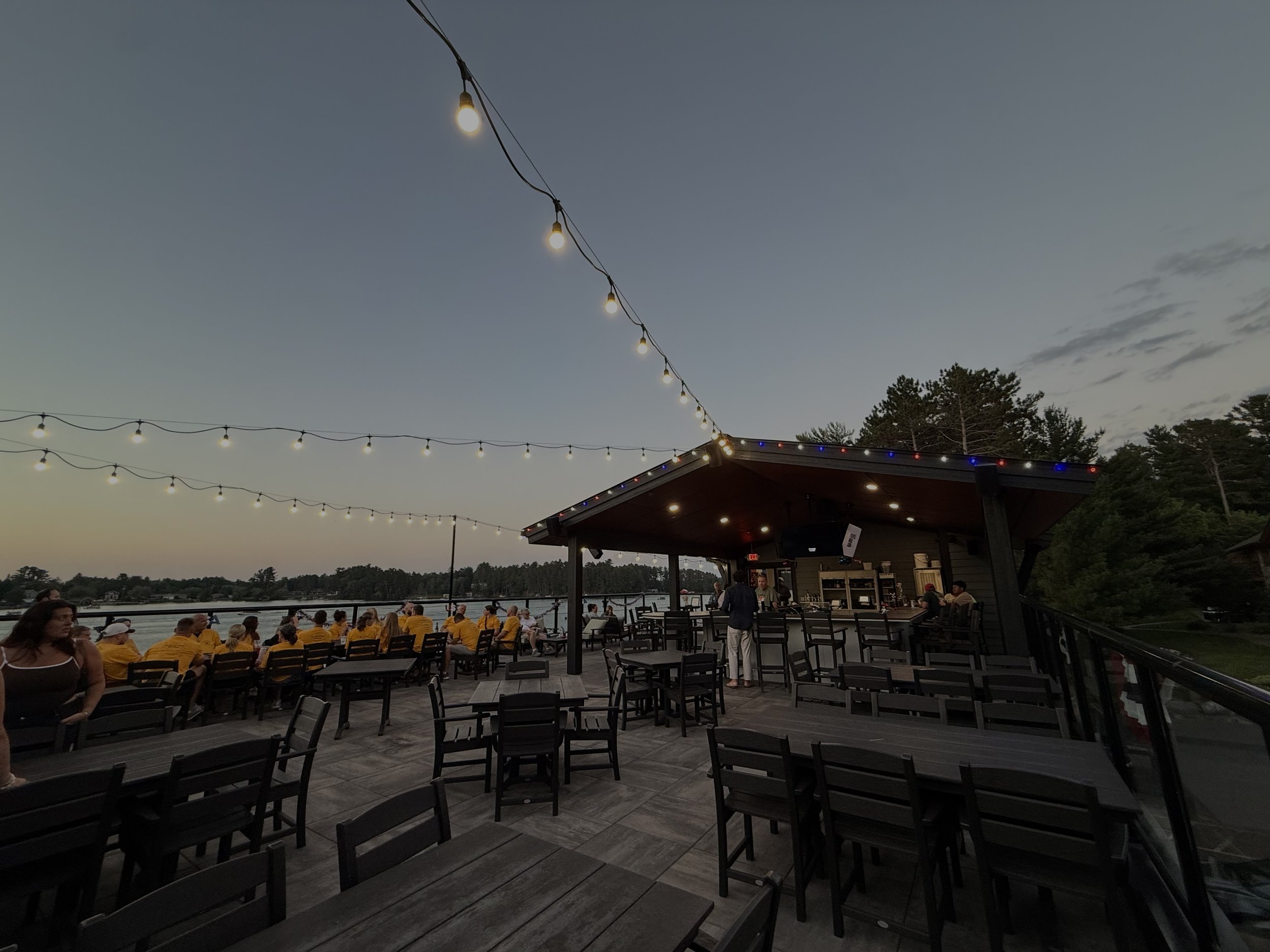 An outdoor dining area at dusk near a body of water with string lights overhead. There are many people sitting at tables, some wearing yellow shirts, with a lakeside view and trees in the background.