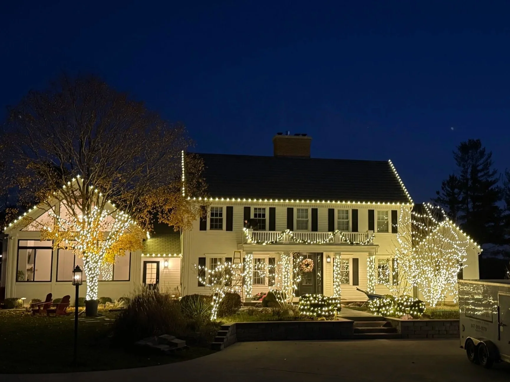 A two-story house decorated with white Christmas lights during nighttime, with trees wrapped in lights and a dark blue sky overhead.