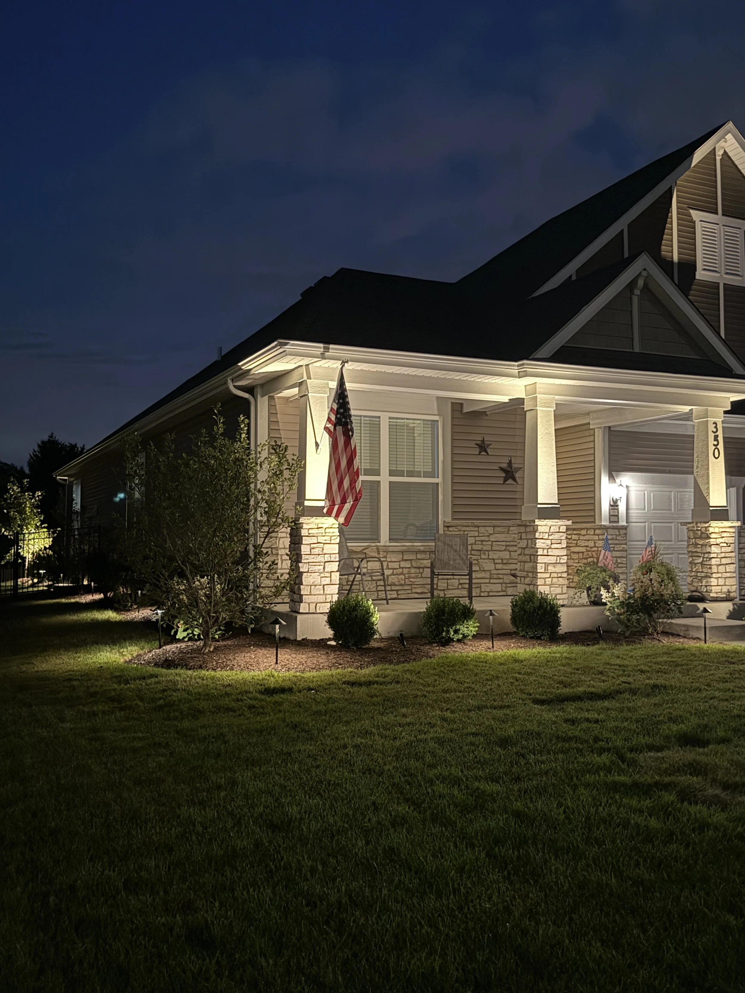 A house exterior at night illuminated by porch lights, with American flags displayed near the entrance, surrounded by a well-maintained lawn and garden.
