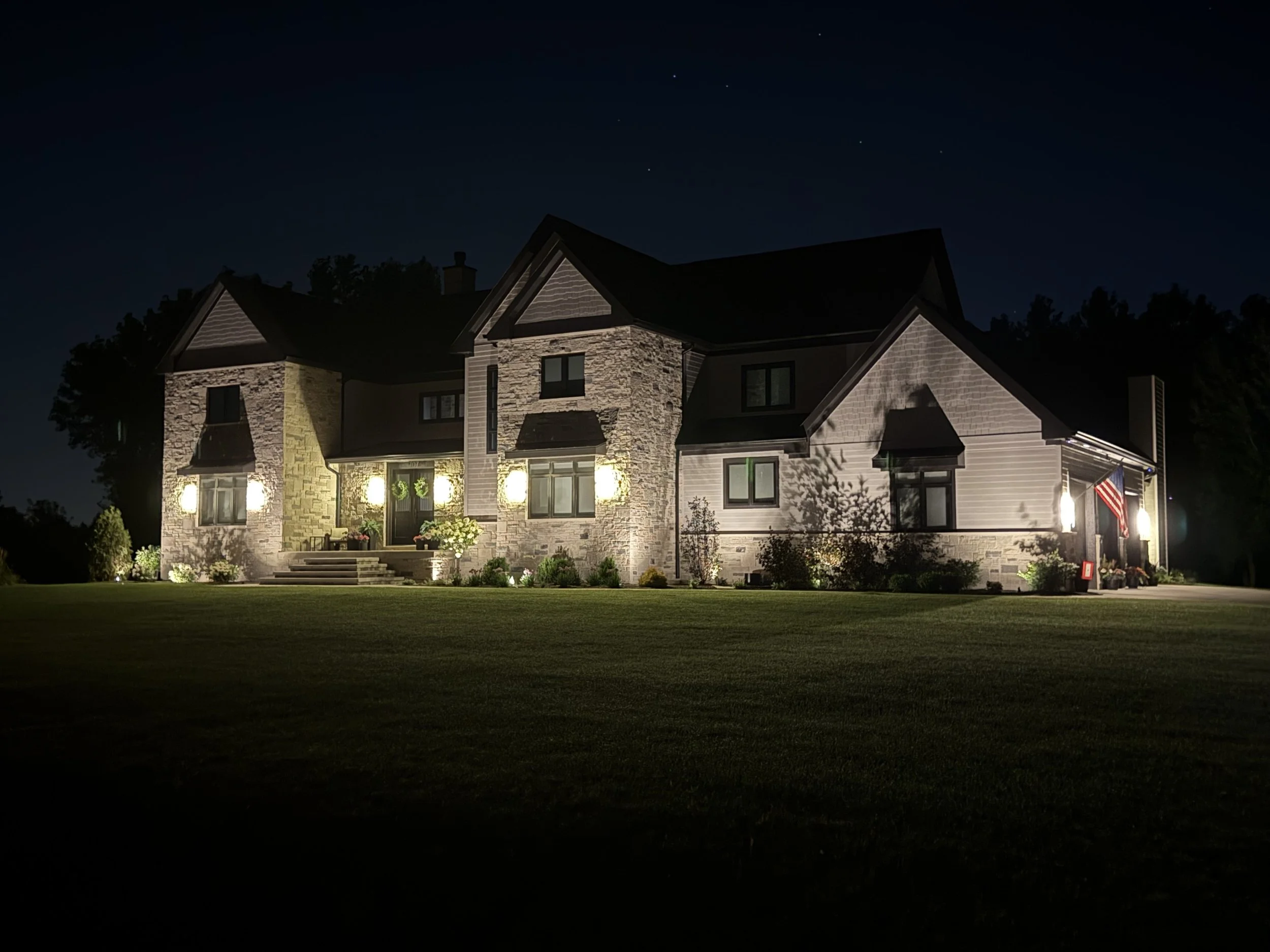 A large house at night with exterior lighting illuminating the front yard and facade. The house has stone and siding exterior, multiple gabled roofs, and an American flag hanging near the porch.