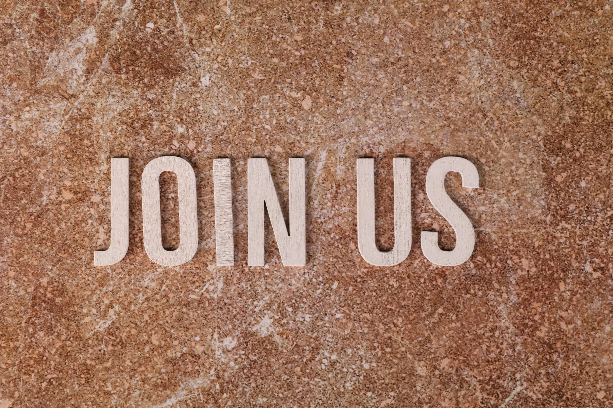 Wooden letters spelling 'JOIN US' placed on a speckled brown surface.
