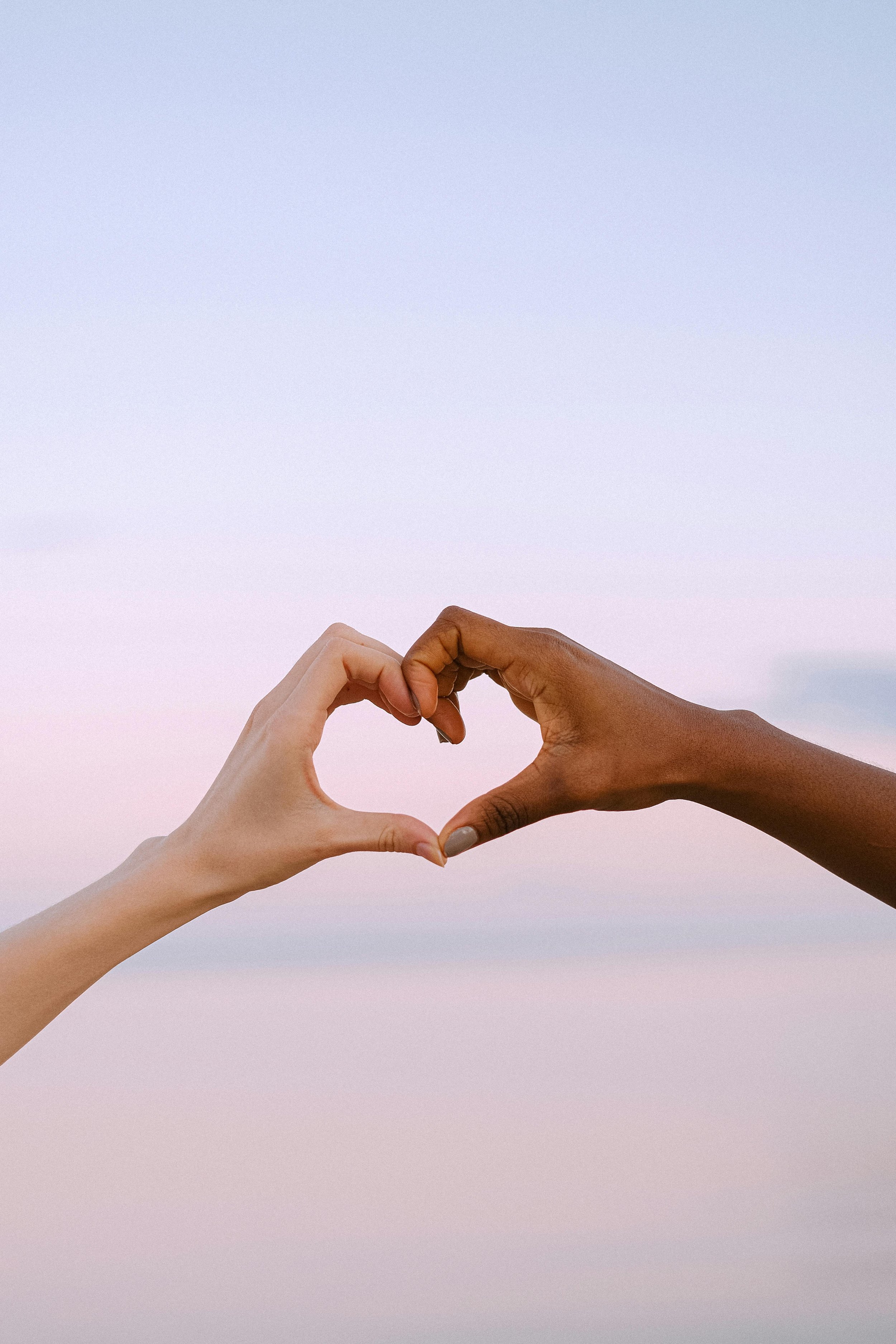 Two hands, one light-skinned and one dark-skinned, forming a heart shape against a pastel sky.