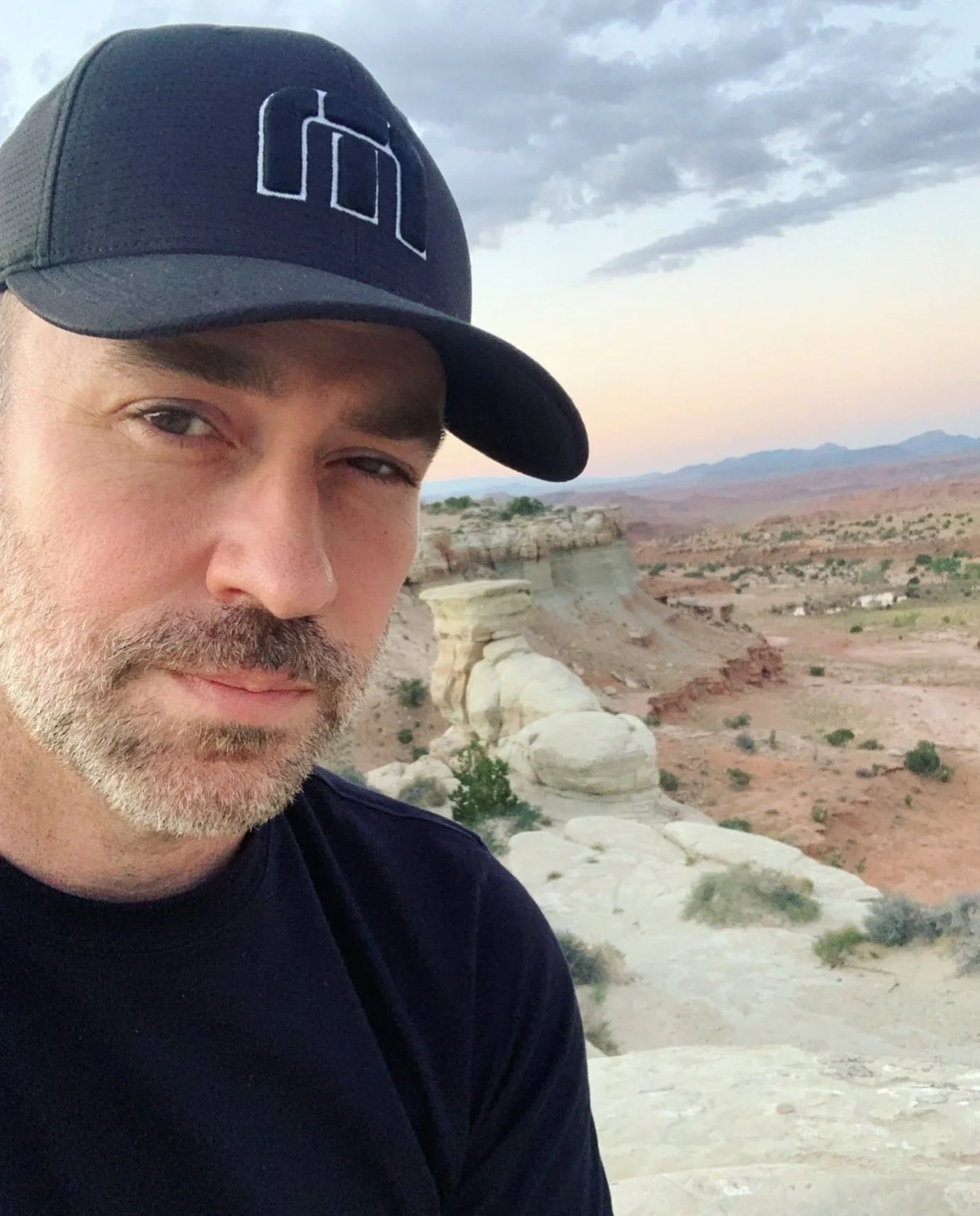 A man with a beard wearing a black cap and black shirt taking a selfie in a desert landscape with rocky formations, sparse vegetation, and distant mountains under a cloudy sky.