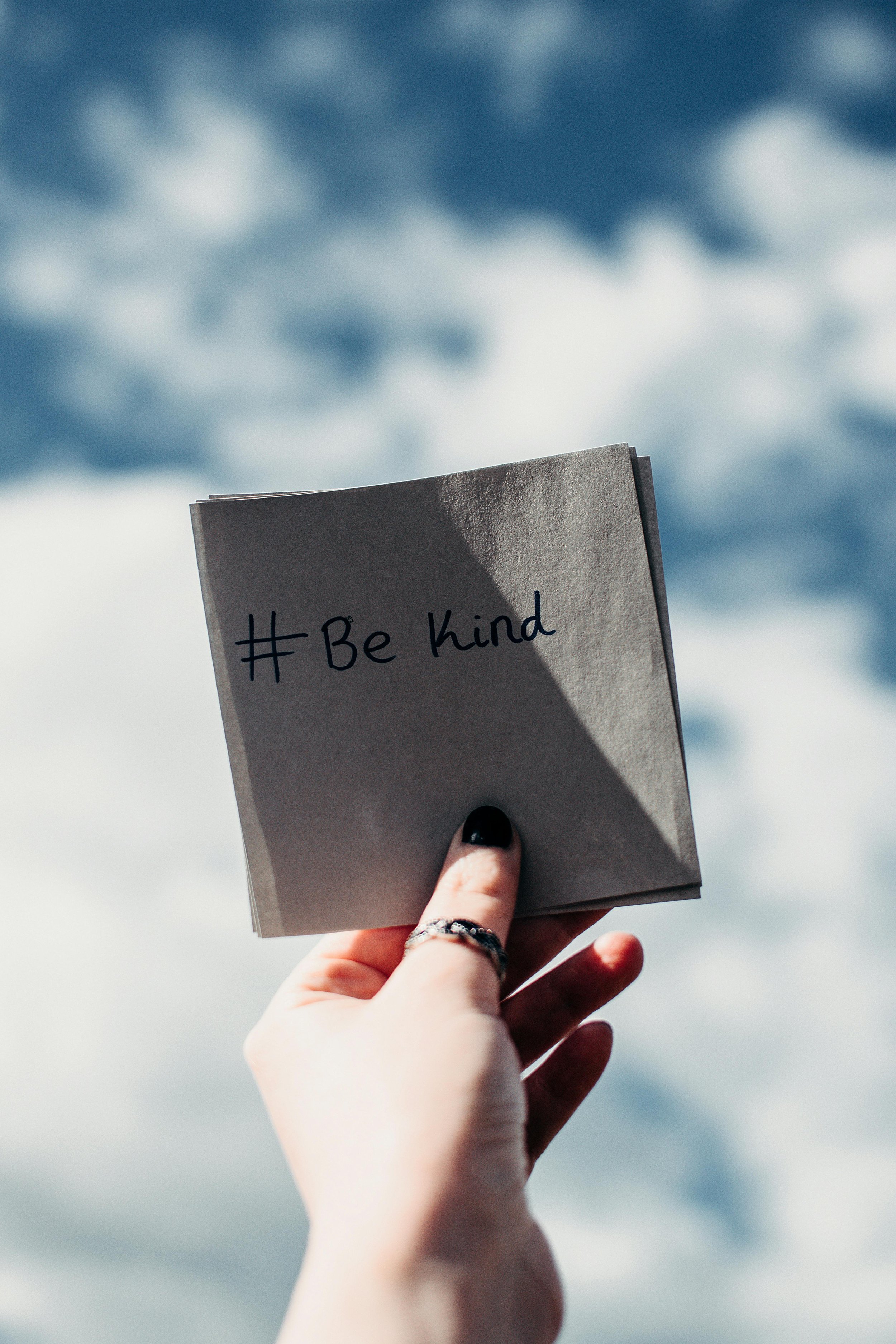 A hand holding a gray card with #BeKind written on it against a sky background with clouds.