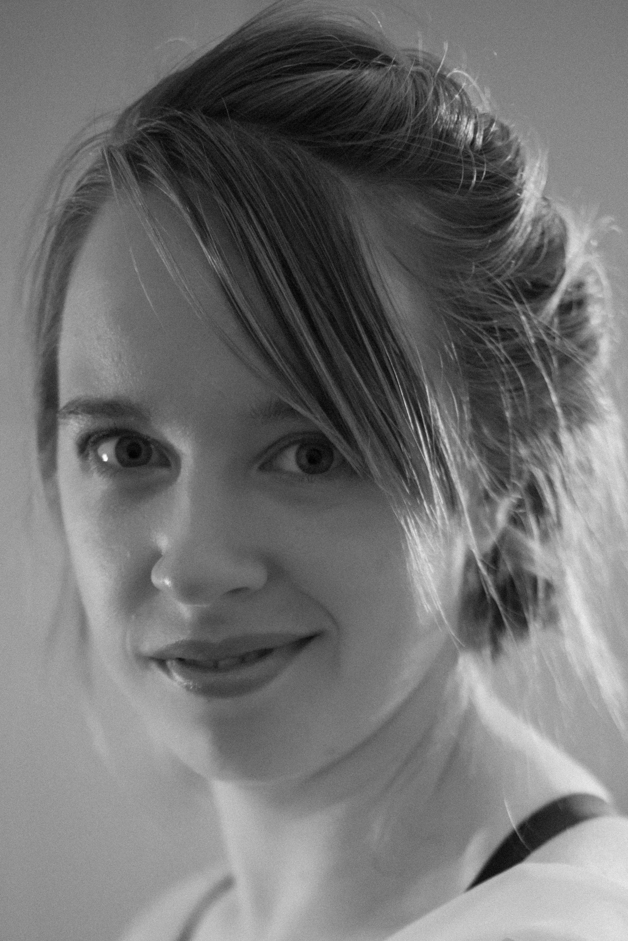 A close-up black-and-white photograph of a woman's face with her hair partially covering her forehead and eyes, showing her nose and lips, with soft lighting and a blurred background.