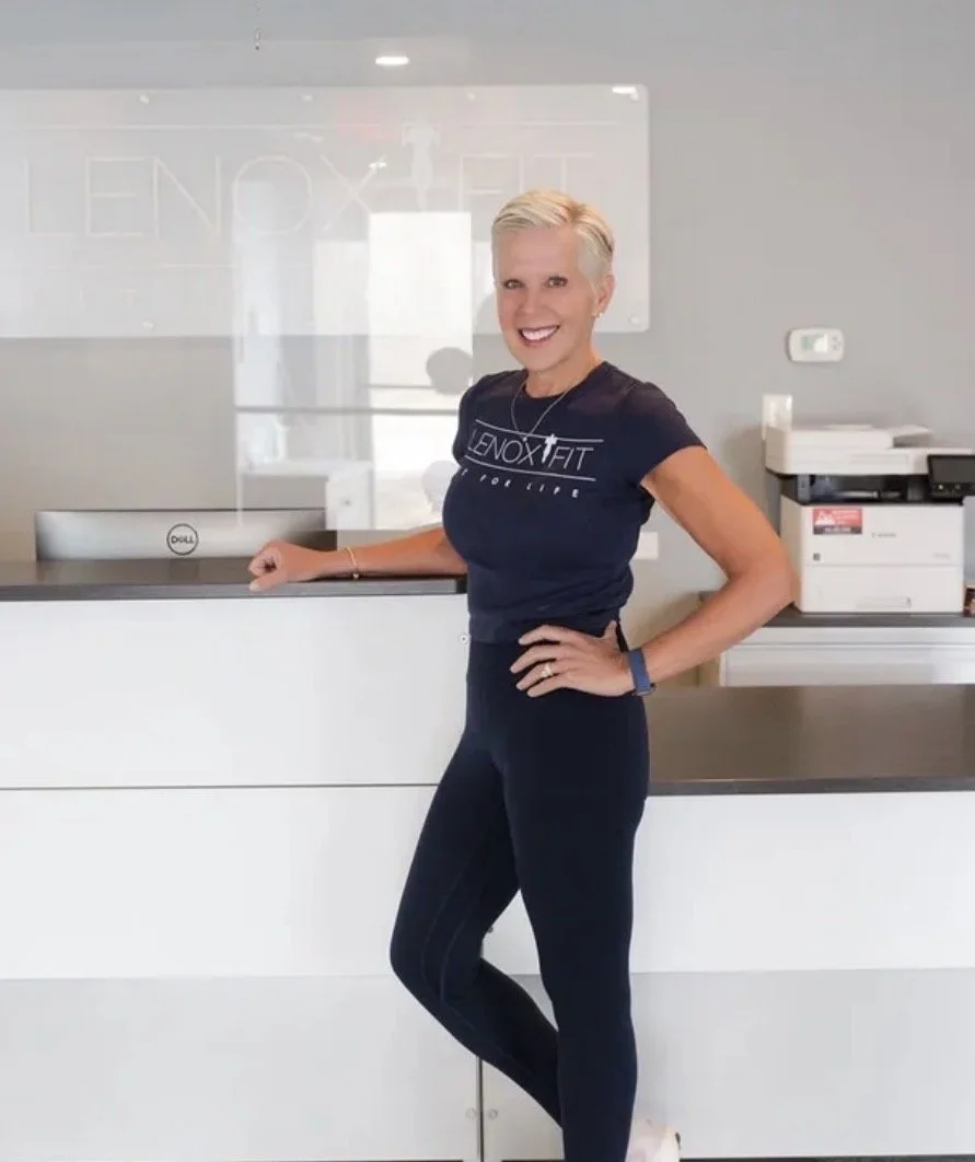 A happy client smiling and posing in front of a reception desk, wearing a black "LENOX FIT" t-shirt and sports leggings. This happy client has worked with us since 2024.