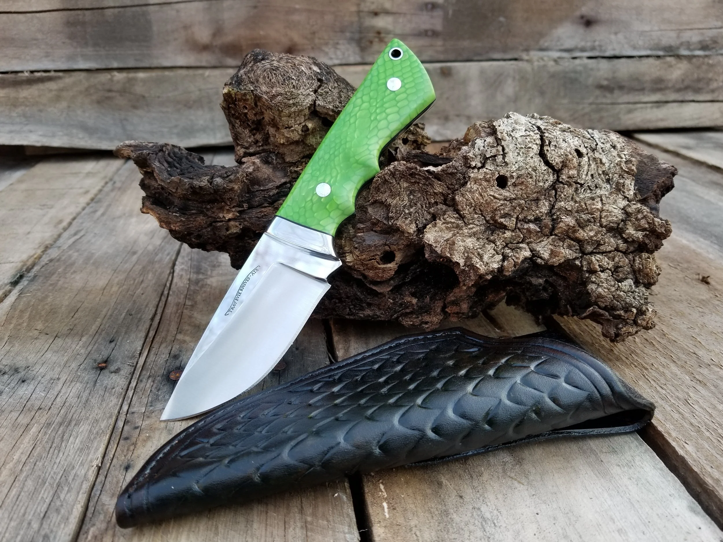 A fixed-blade knife with a green textured handle, a black leather sheath, and a piece of bark and wood placed together in a rustic setting.