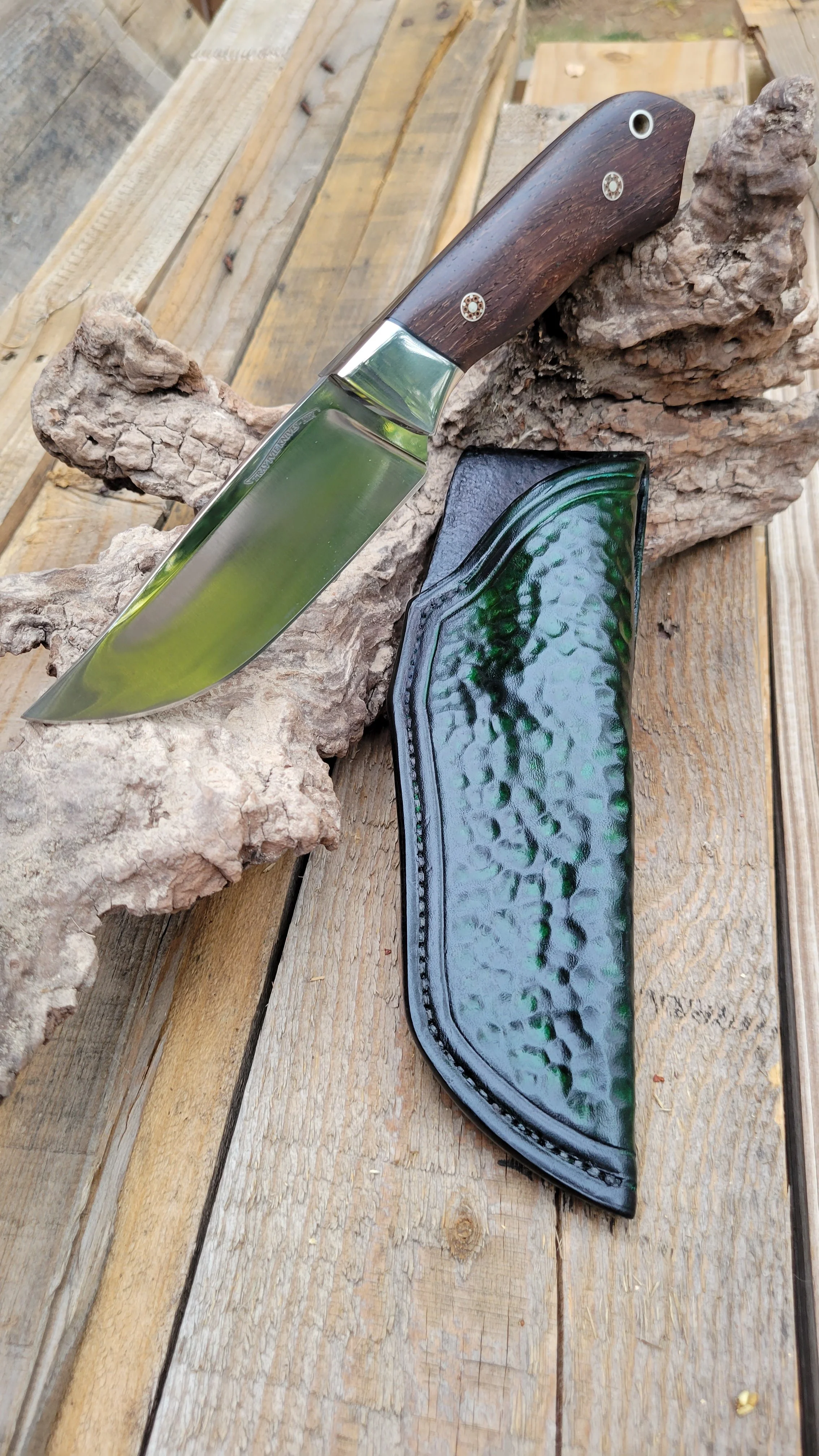 A fixed-blade hunting knife with a wooden handle resting on a piece of driftwood, alongside a black textured leather sheath, on a wooden surface.