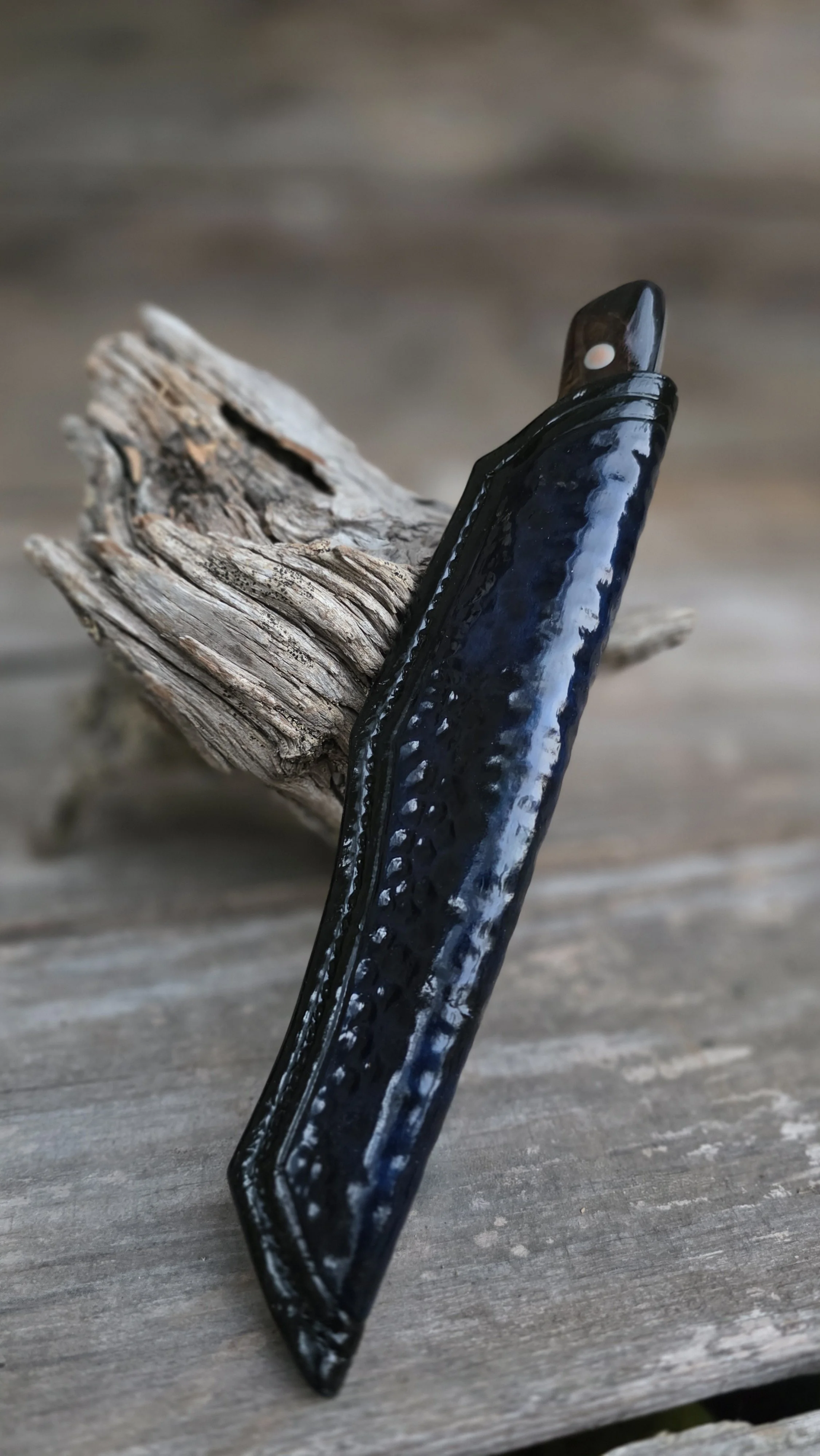 Close-up of a black-handled straight razor with a textured surface, resting against a piece of weathered driftwood on a wooden surface.