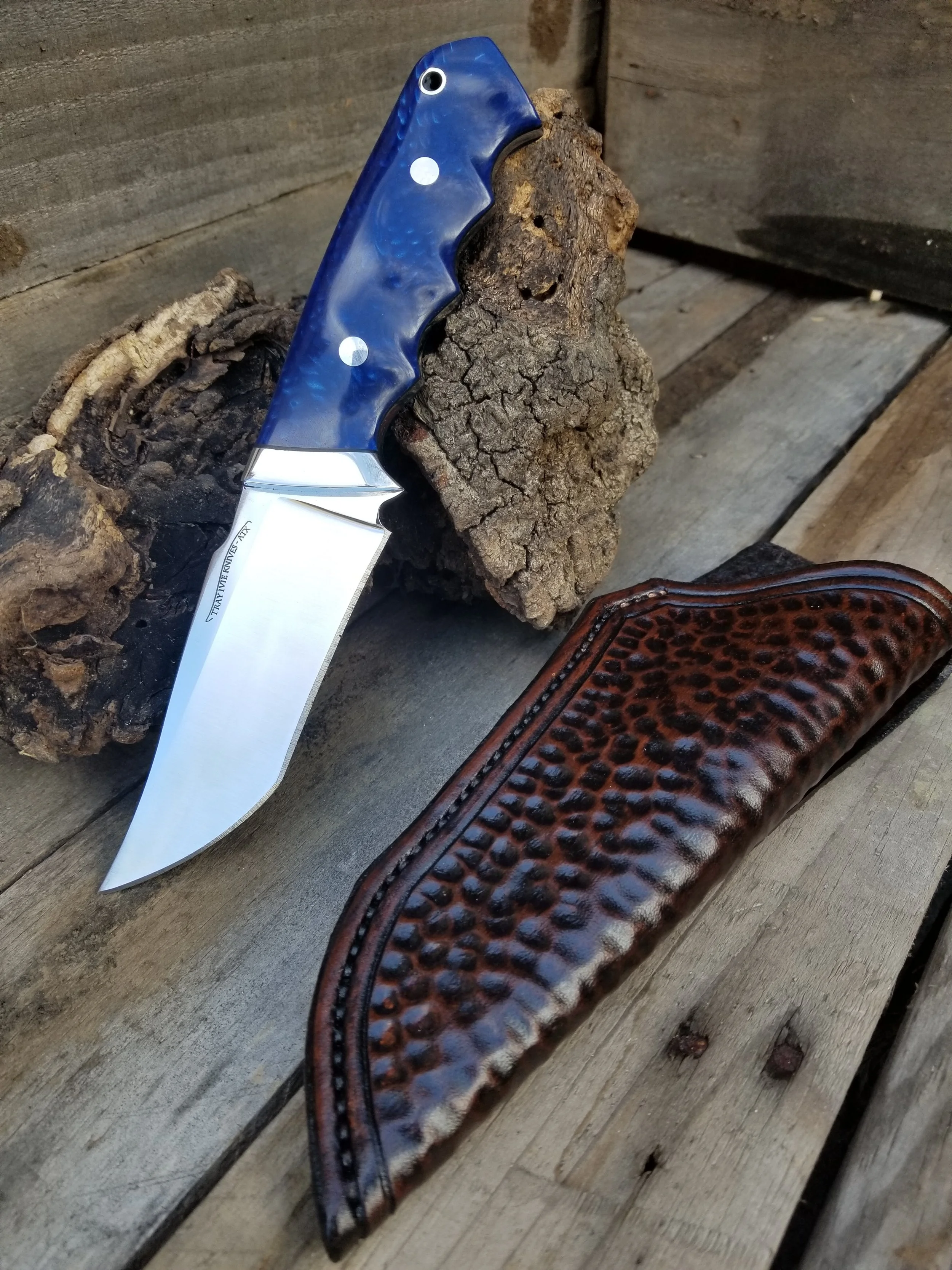 A fixed-blade hunting knife with a blue handle and a polished metal blade, resting on a piece of bark, with a leather sheath nearby on a wooden surface.