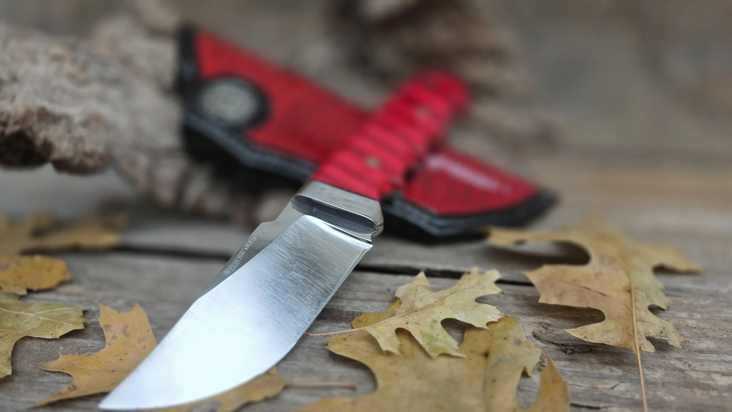 A folding pocket knife with a red handle and stainless steel blade resting on a weathered wooden surface surrounded by yellow autumn leaves.
