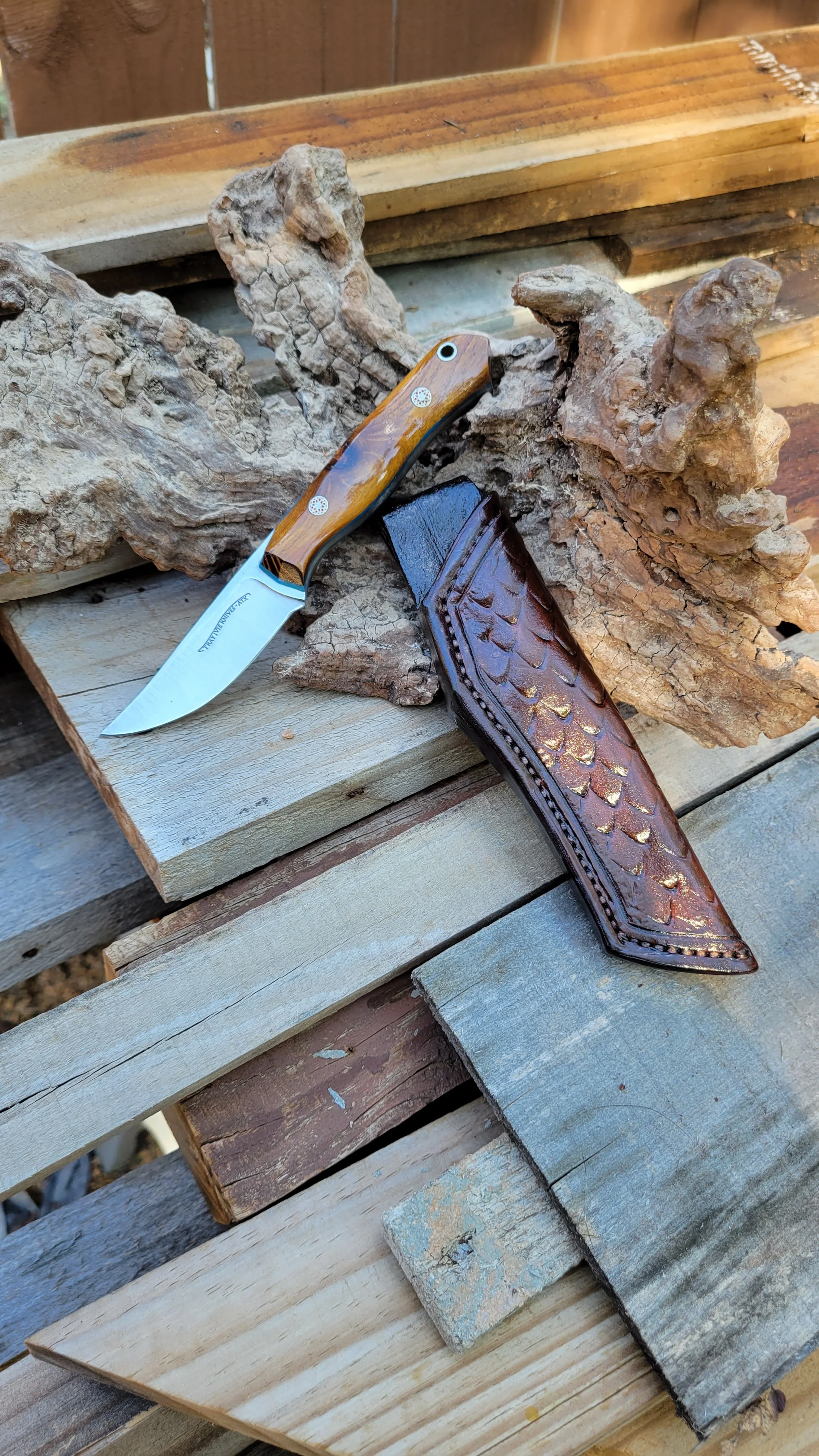 A pocket knife with a wooden handle and a sharp blade resting on a piece of driftwood, next to a leather sheath with a scale pattern, on a wooden surface outdoors.