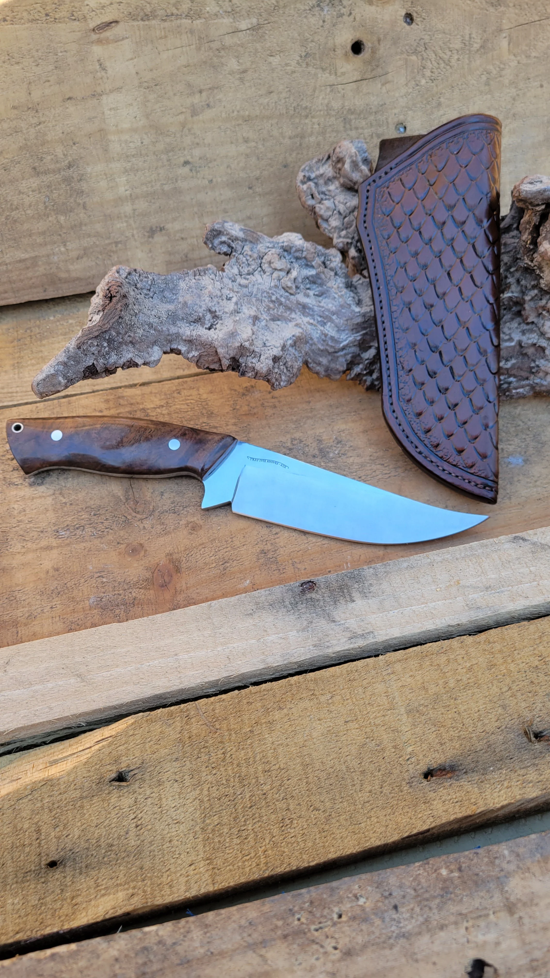 A fixed-blade knife with a wooden handle, a textured leather sheath, a piece of weathered driftwood, and a wooden surface.