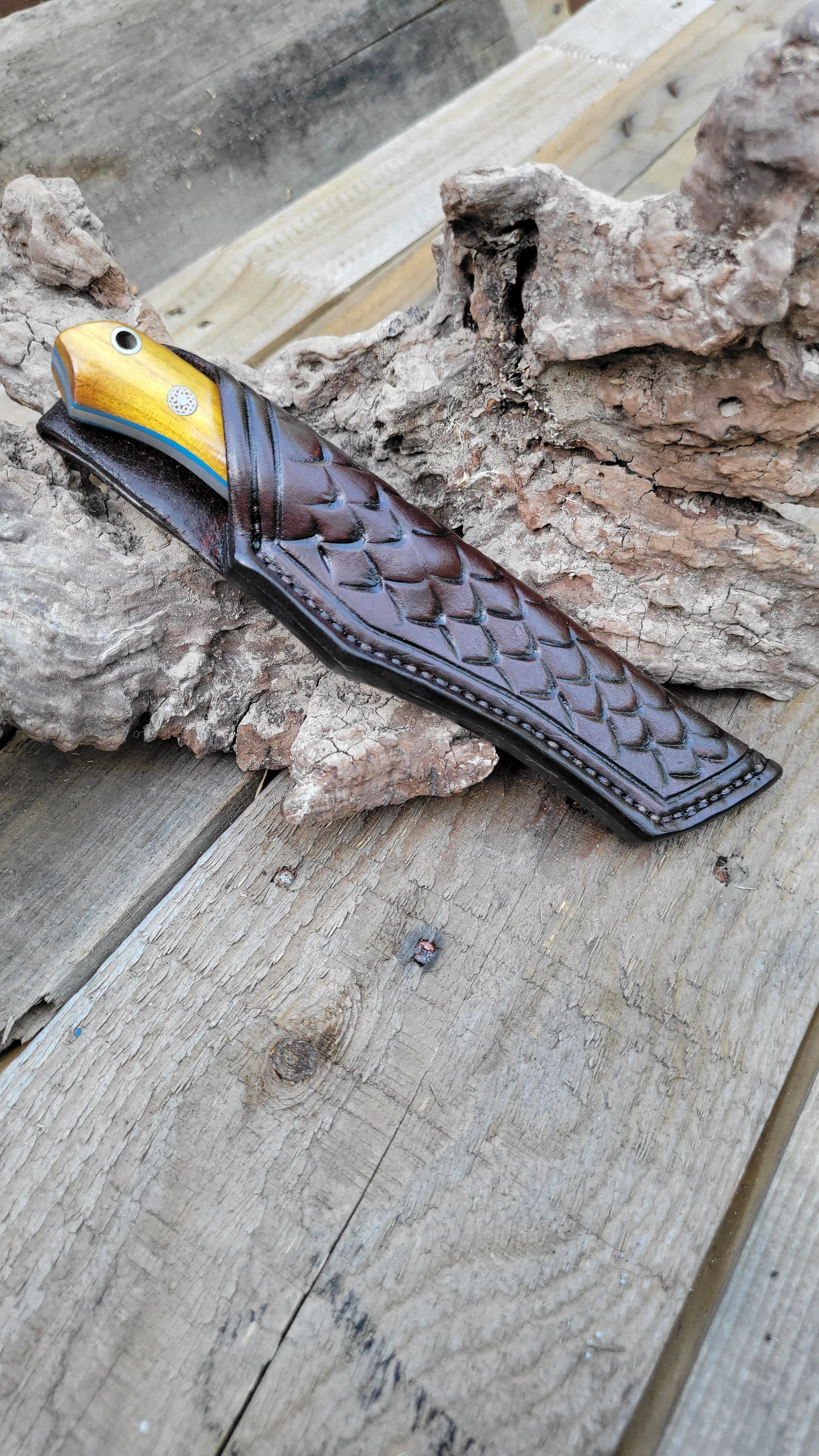 A fish-shaped knife with a yellow and blue handle and a black scaled pattern on the blade, resting on a rock on a wooden surface.