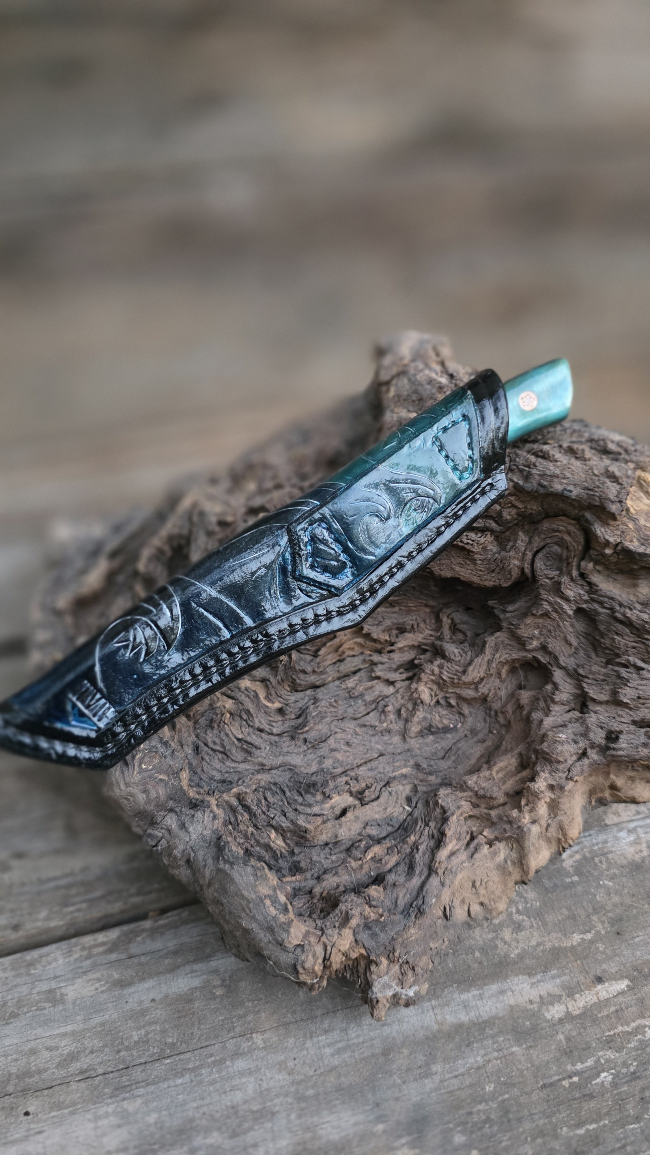 A paisley-patterned knife with a turquoise handle resting on a piece of driftwood on a weathered wooden surface.