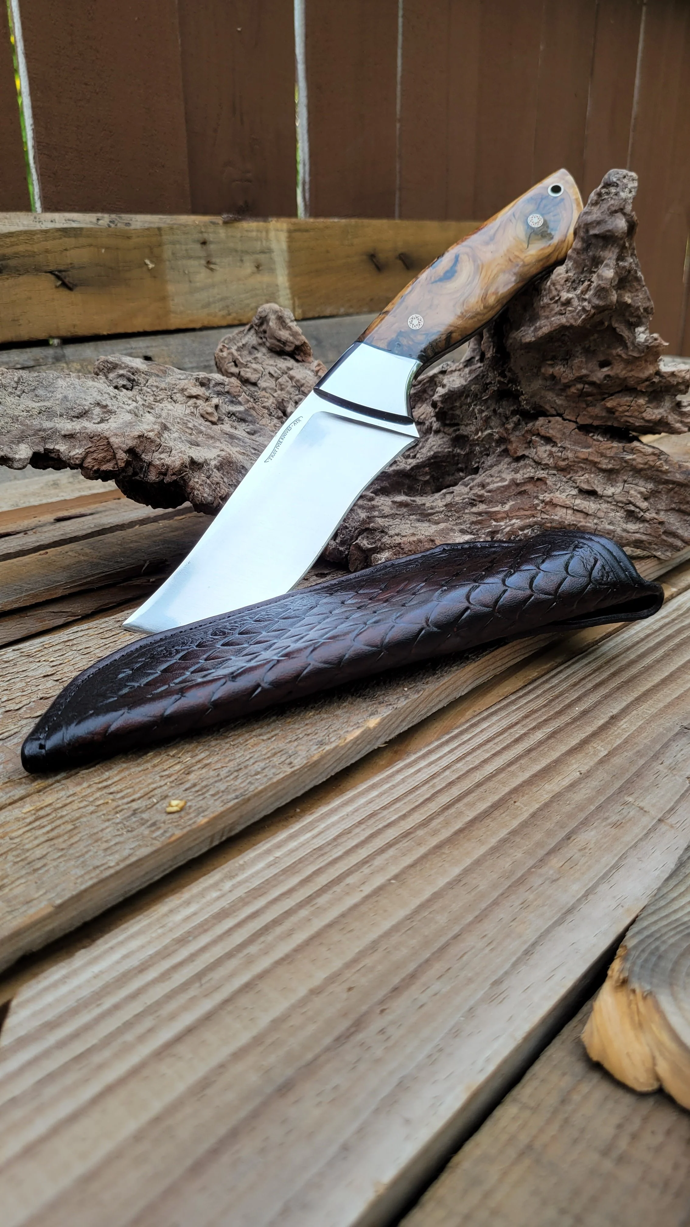 A kitchen knife with a polished blade and a handle made of wood, resting on a wooden surface with a leather sheath nearby.