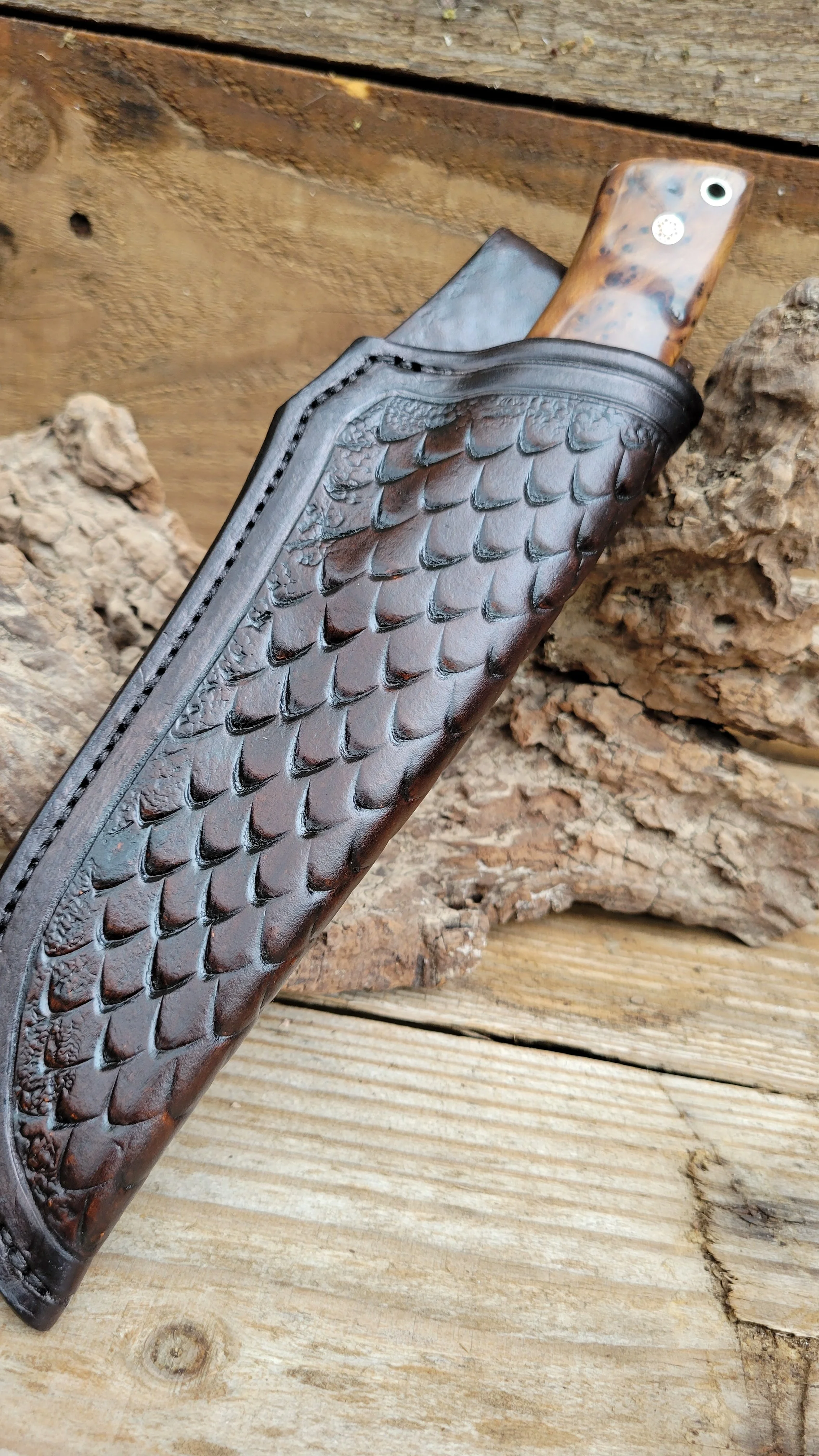 Close-up of a handcrafted knife handle made of burl wood, resting in a leather sheath with a textured, scale-like pattern, on a wooden surface with wood logs in the background.
