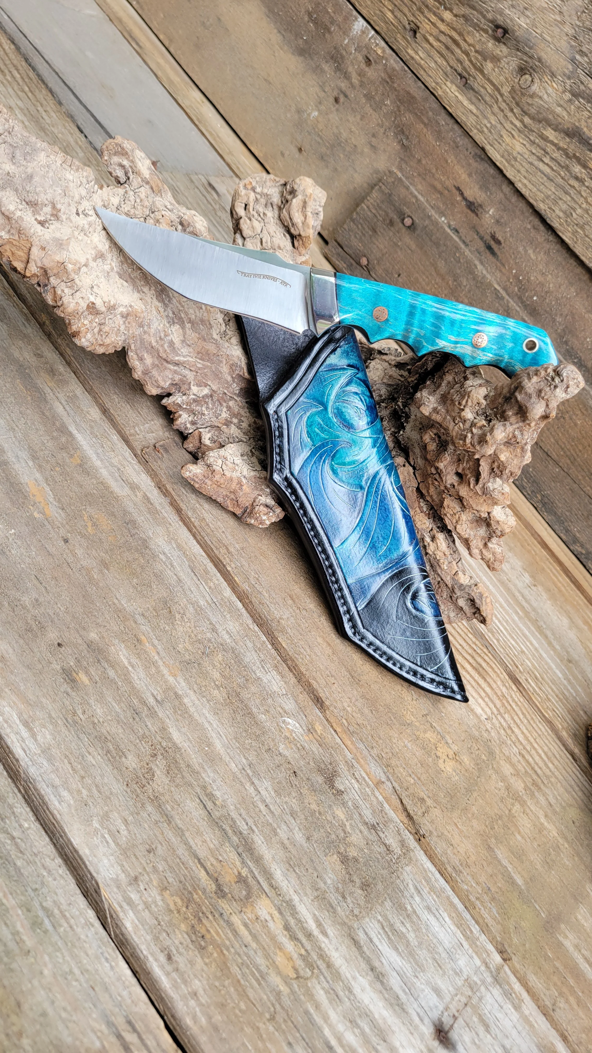 A colorful folding pocket knife with a blade and a handle in shades of turquoise and black, resting on textured bark and a rustic wooden surface.