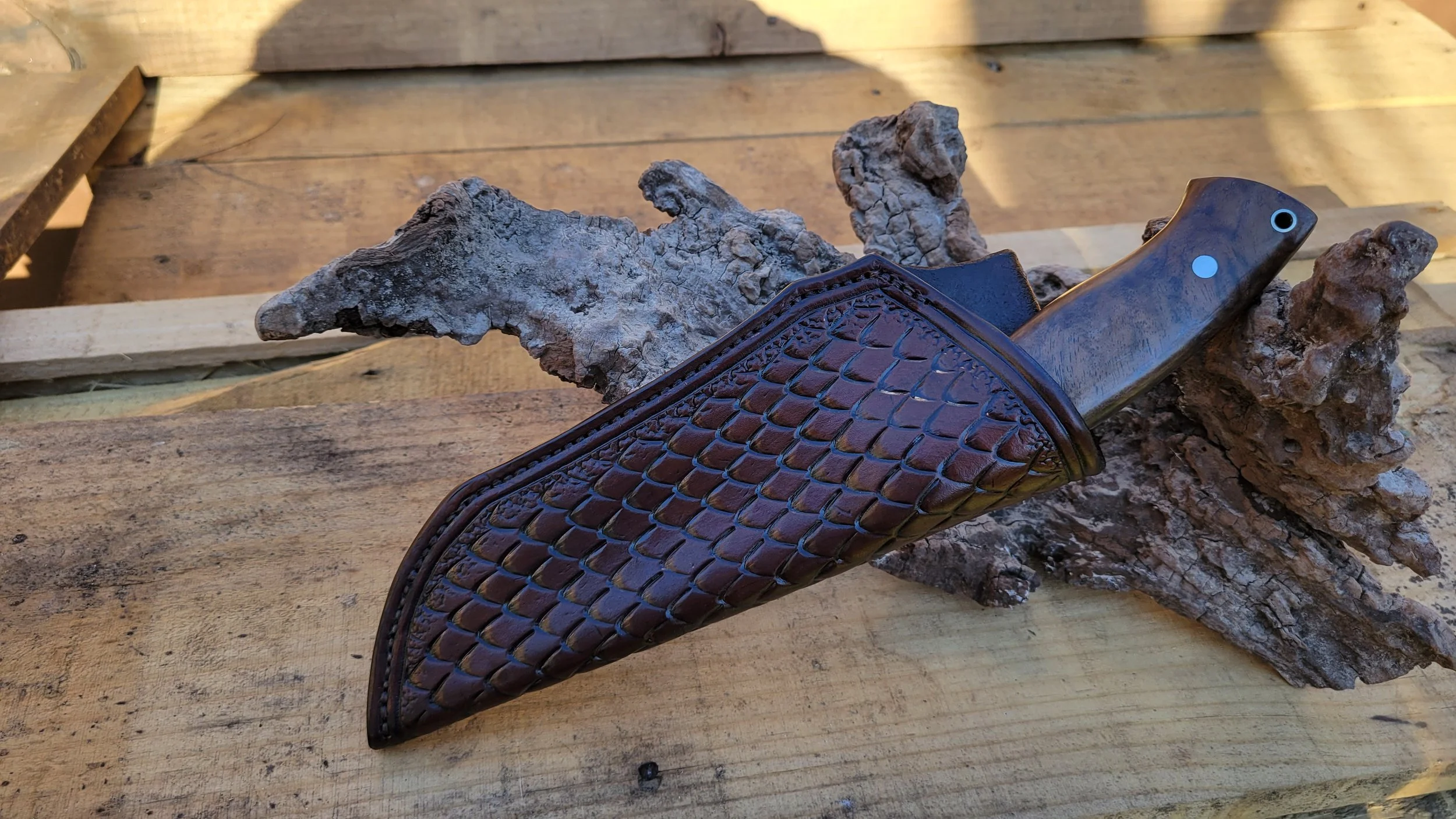 A hunting knife with a wood handle and a leather sheath featuring a scaled pattern, resting on a piece of bark and a wooden surface.