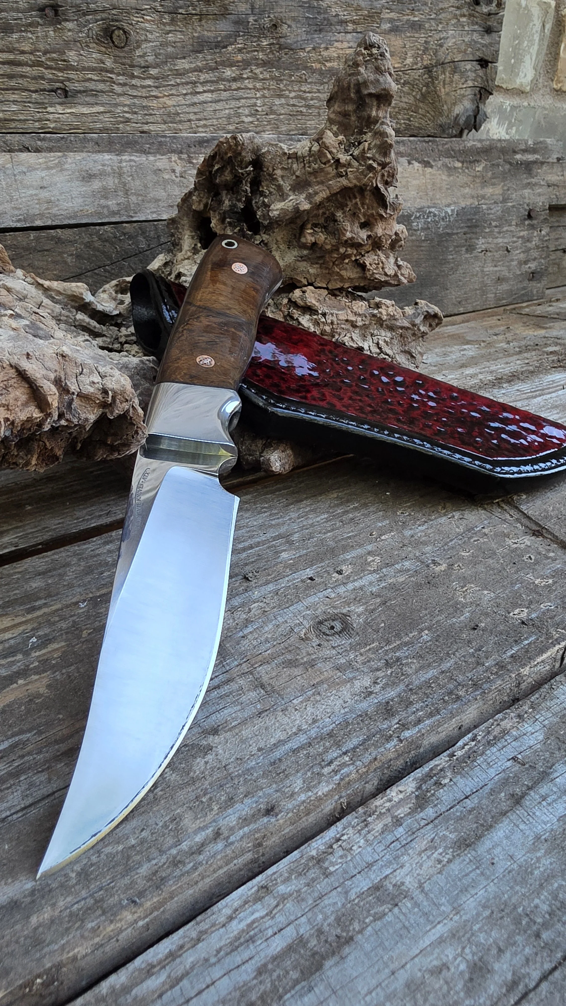 A fixed-blade knife with a polished wood handle resting on a weathered wooden surface, with a red and black sheath leaning against a piece of driftwood.
