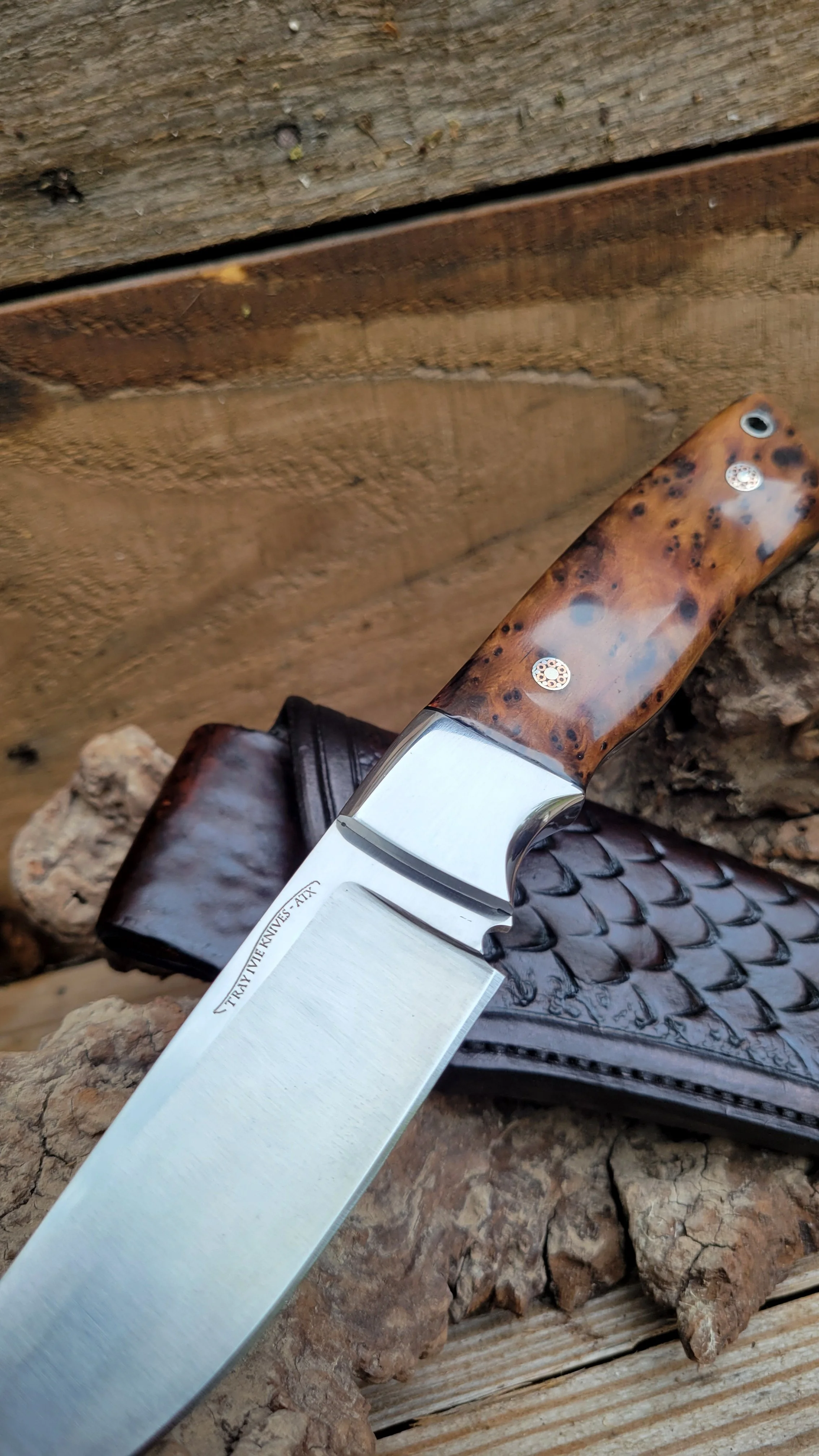 Close-up of a fixed-blade knife with a wooden handle and a slightly curved blade, resting on a textured surface with rocks and a leather sheath.