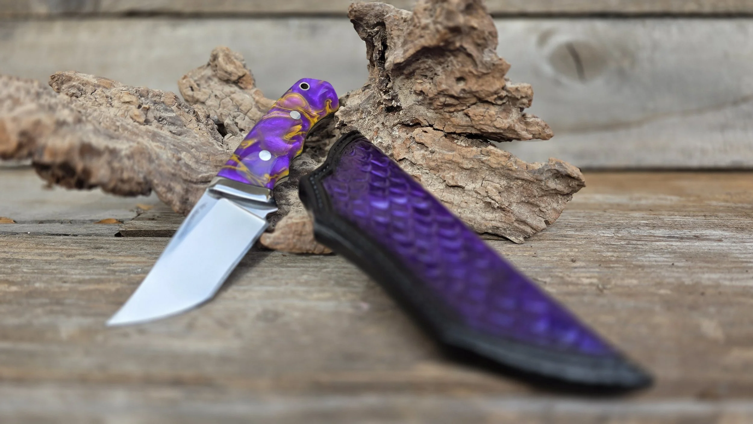 A folding knife with a purple and yellow swirl handle and a metal blade, resting on a piece of textured purple material and a piece of bark on a wooden surface.