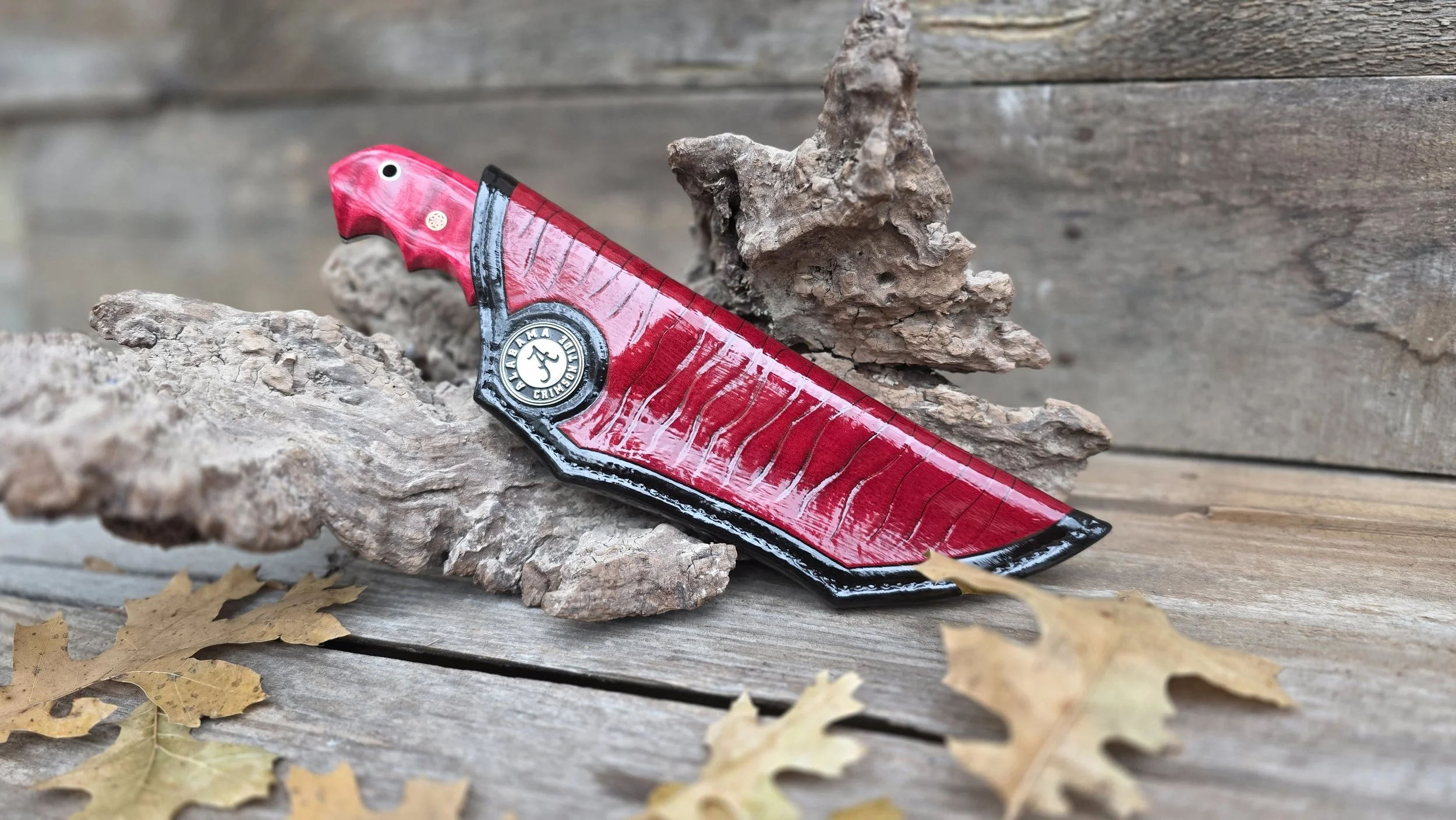 A red and black folding knife with a textured handle, resting on a piece of driftwood on a weathered wooden surface, with dried autumn leaves nearby.