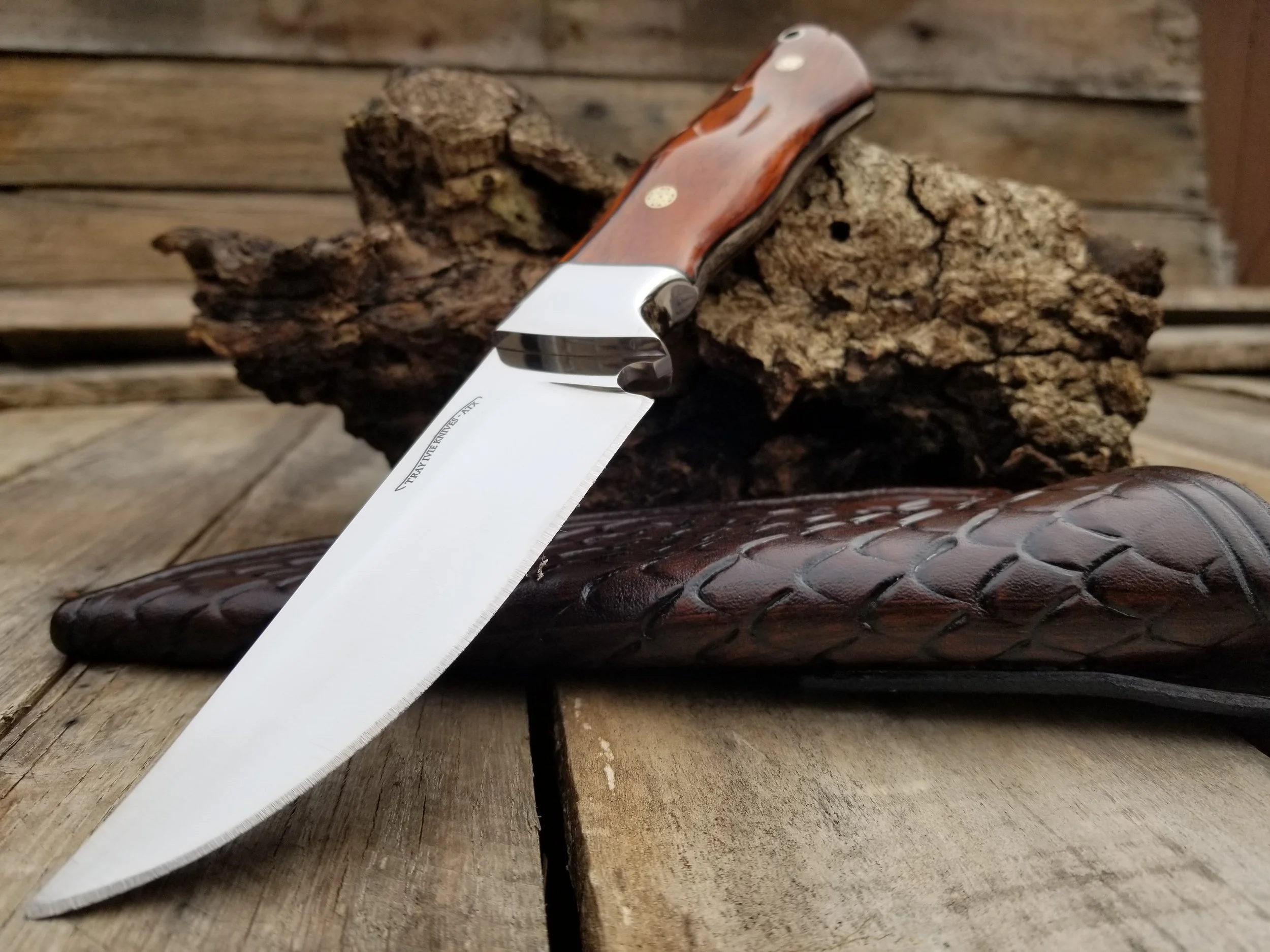 A chef's knife with a wooden handle resting on a piece of wood and a leather glove, with a piece of bark in the background.