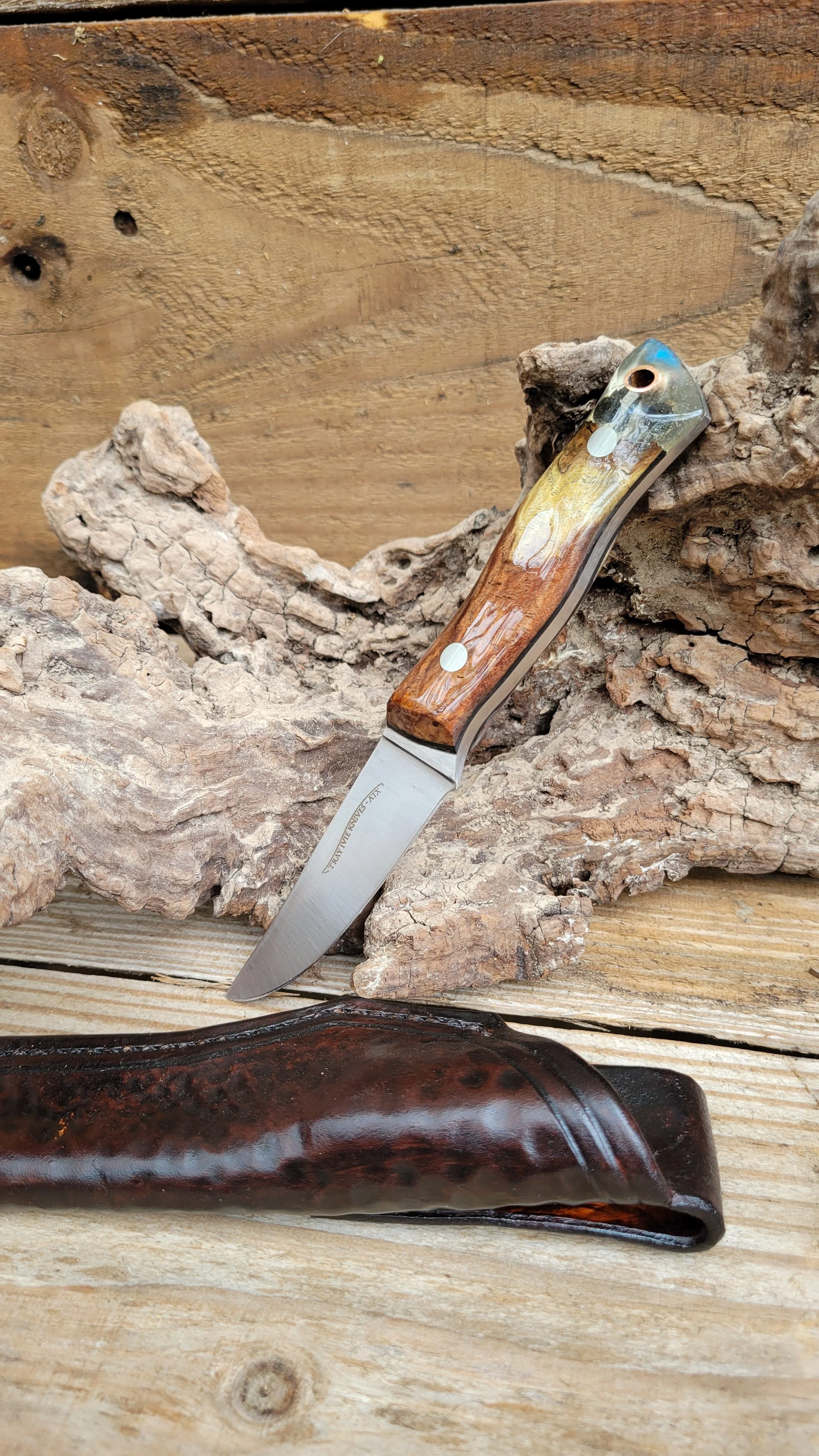 A small folding knife with a wooden handle, partially opened, resting on a piece of rough bark. A leather sheath is in the foreground on a wooden surface, and a wooden backdrop is in the background.