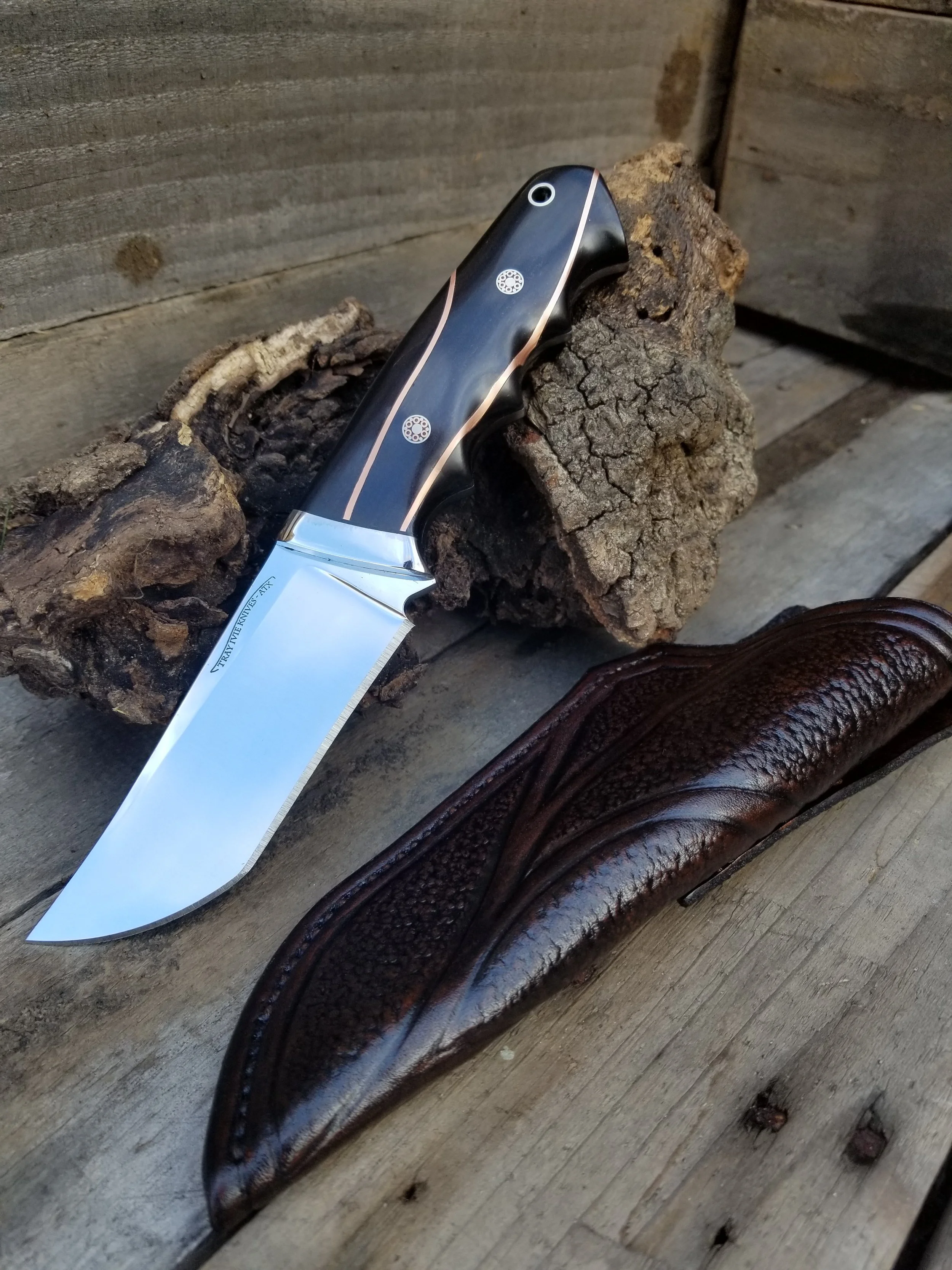 A large fixed-blade outdoor knife resting on a piece of wood, with a dark leather sheath nearby, on a wooden surface.