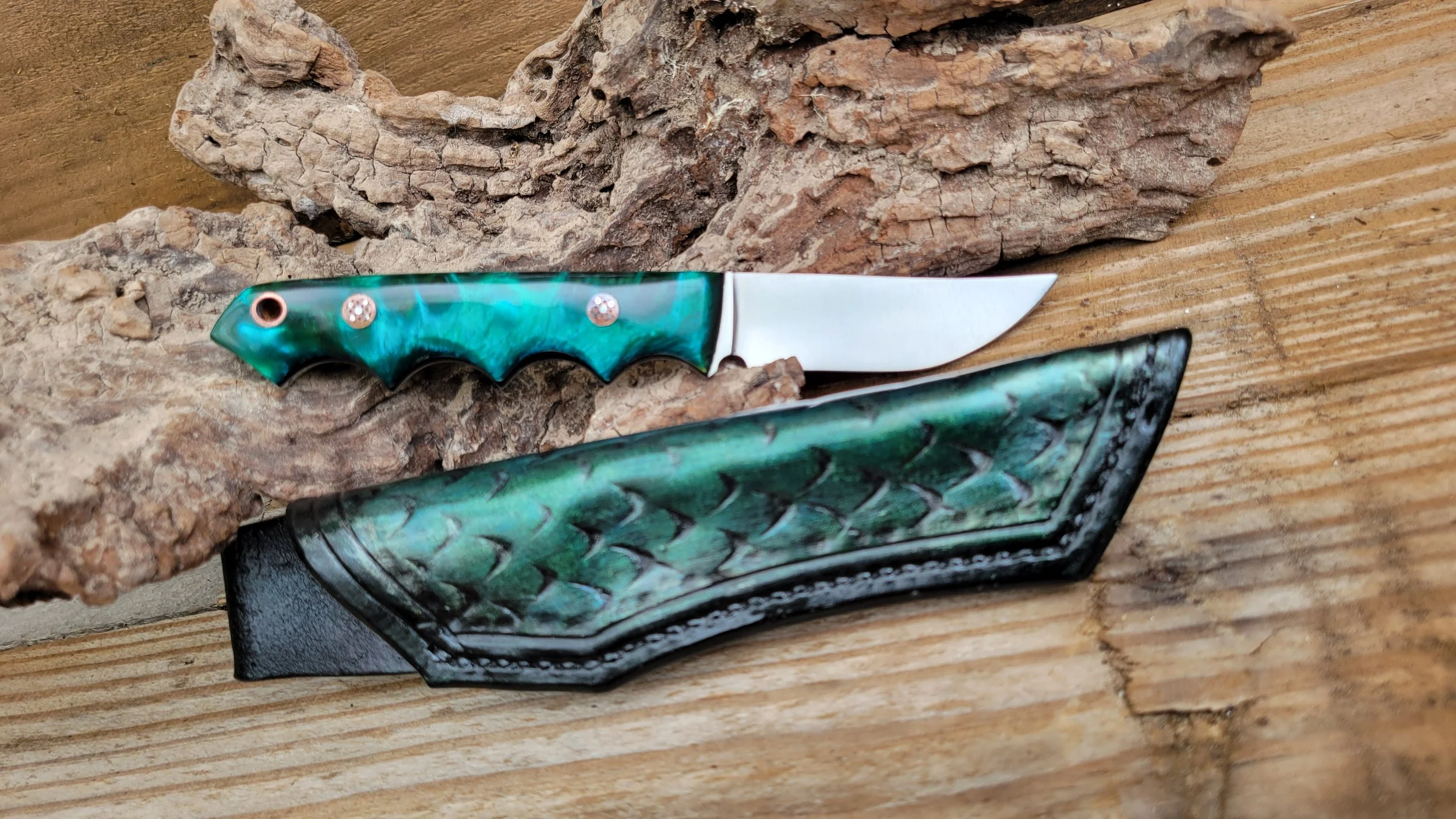 A folding pocket knife with a greenish-blue handle featuring gemstone accents, displayed on a wooden surface with a piece of driftwood.