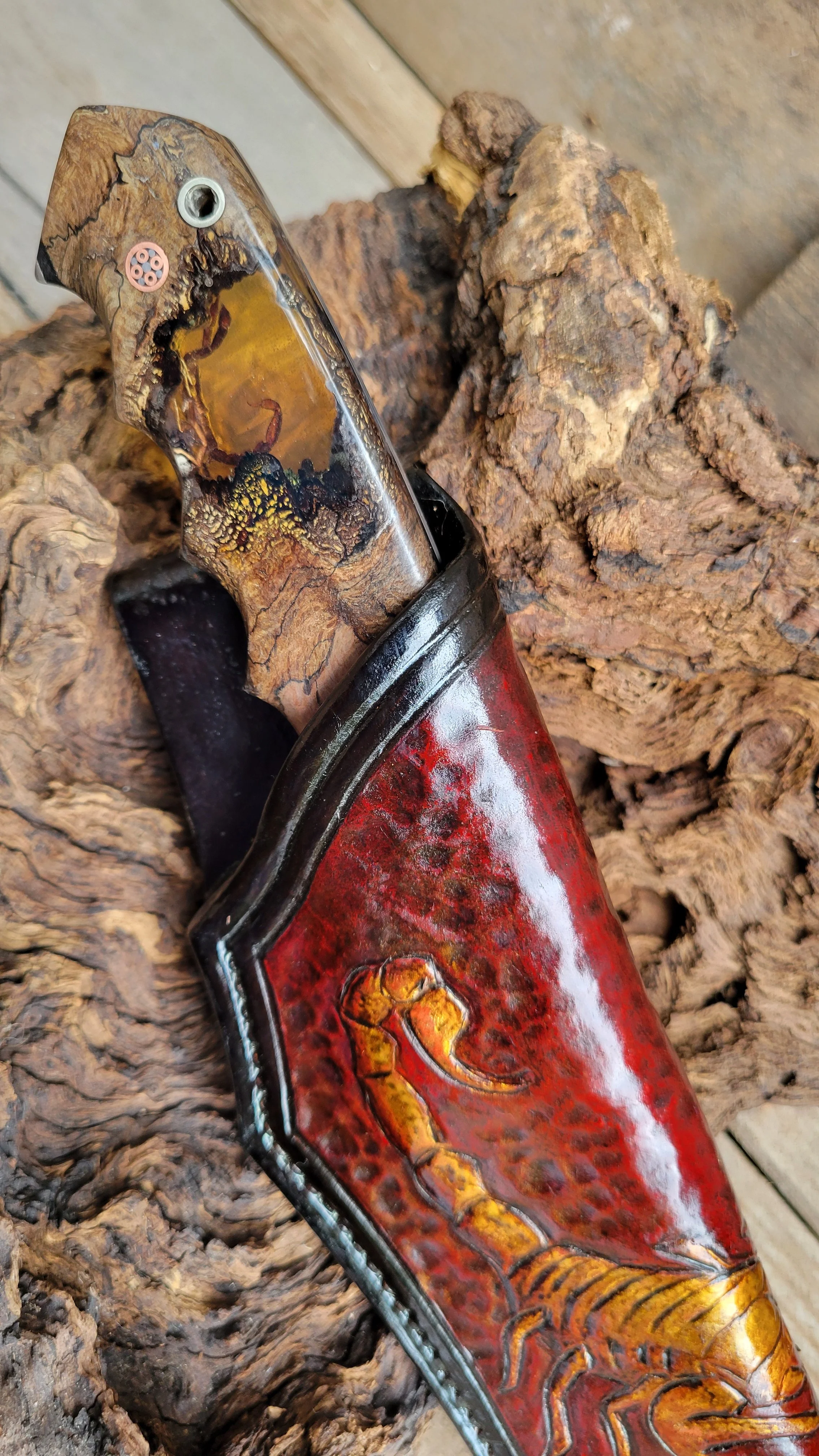 A folding knife with a handle made of wood and resin, featuring a scorpion engraved on the red case, resting on a piece of textured wood.