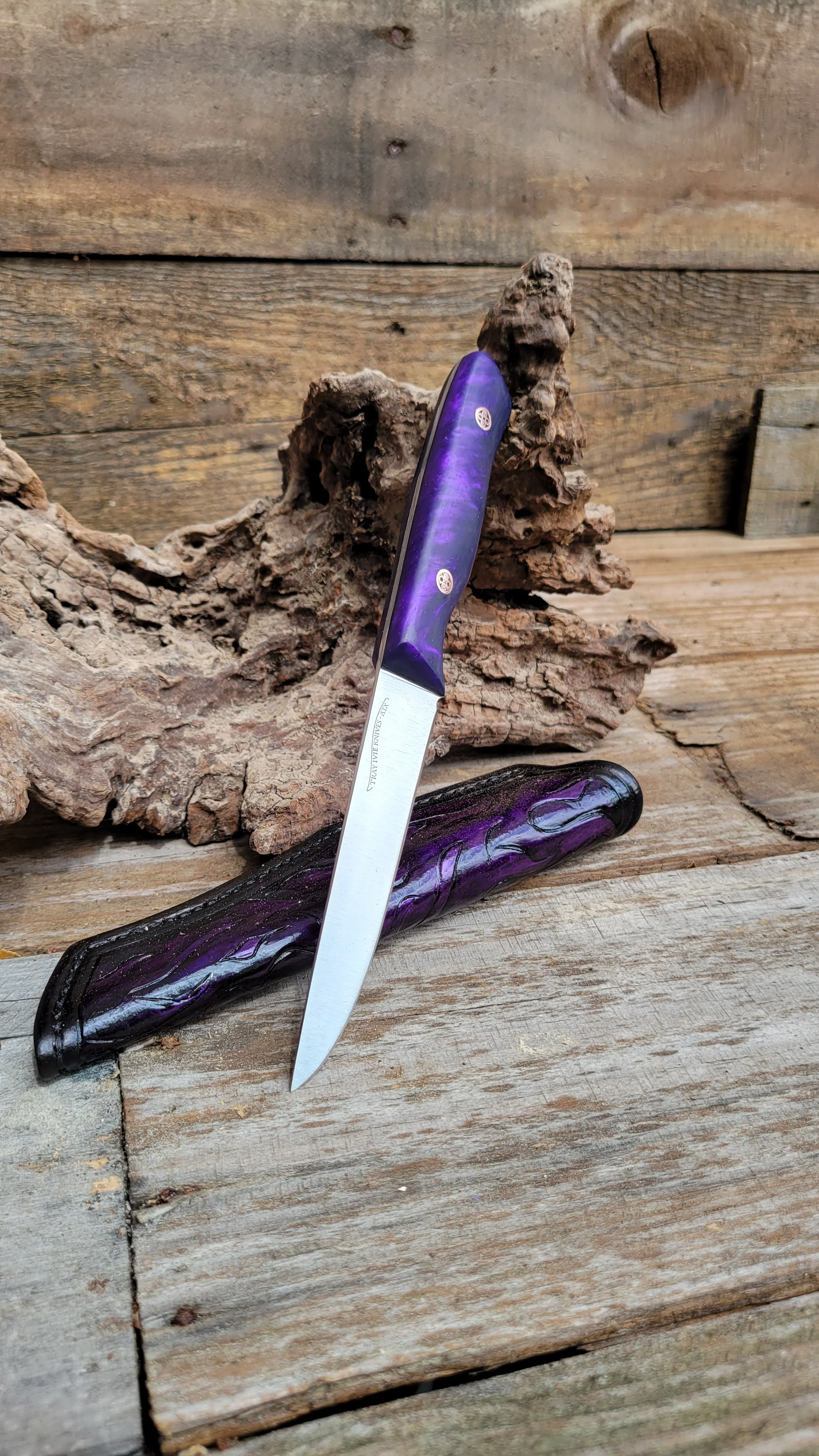 A pocket knife with a purple and black handle resting on a textured black sheath, set on a rustic wooden surface with a piece of driftwood and wooden planks in the background.