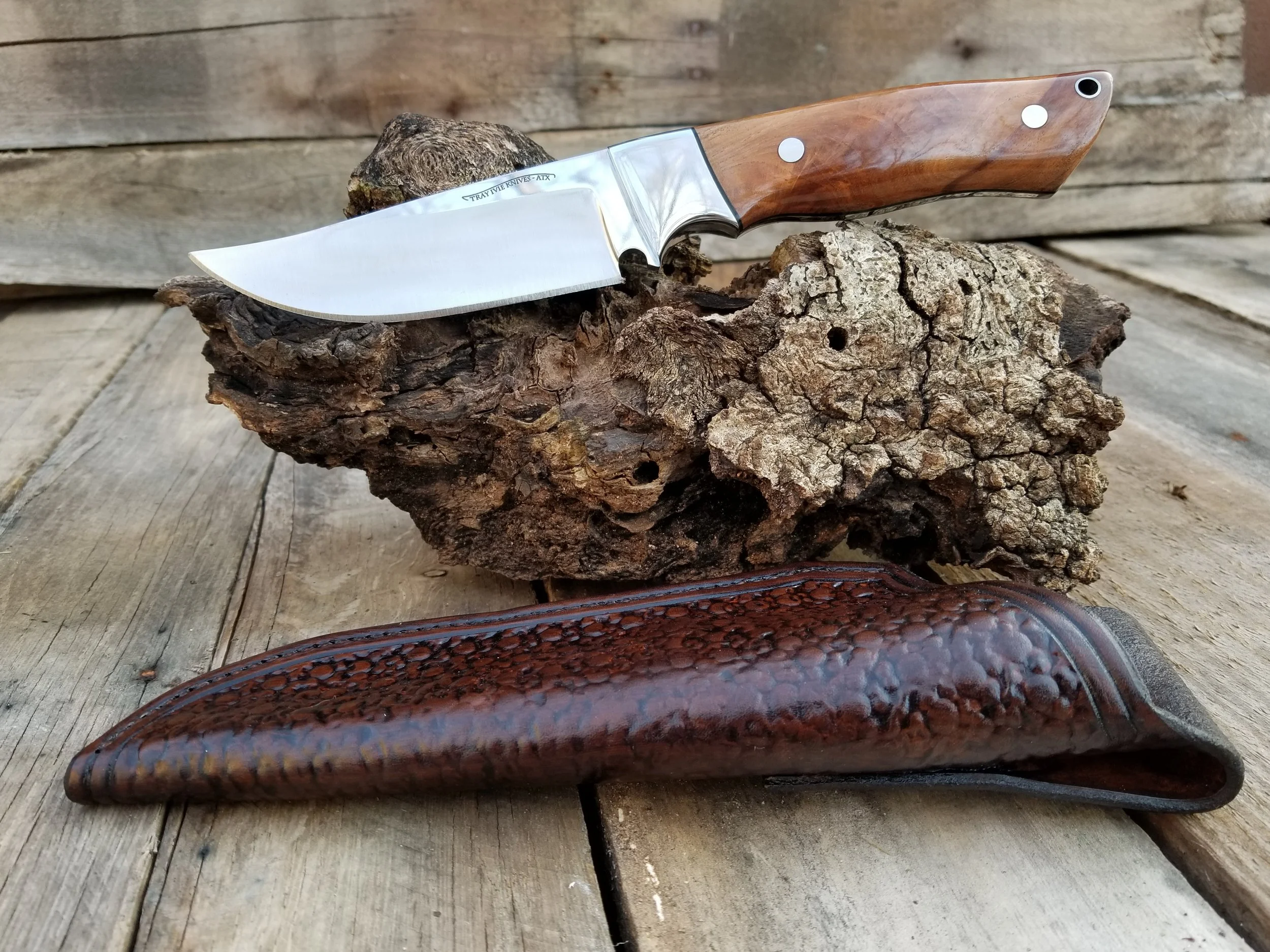 A hunting knife with a wooden handle and a stainless steel blade, resting on a piece of bark with a brown leather sheath below it, all placed on a wooden surface.