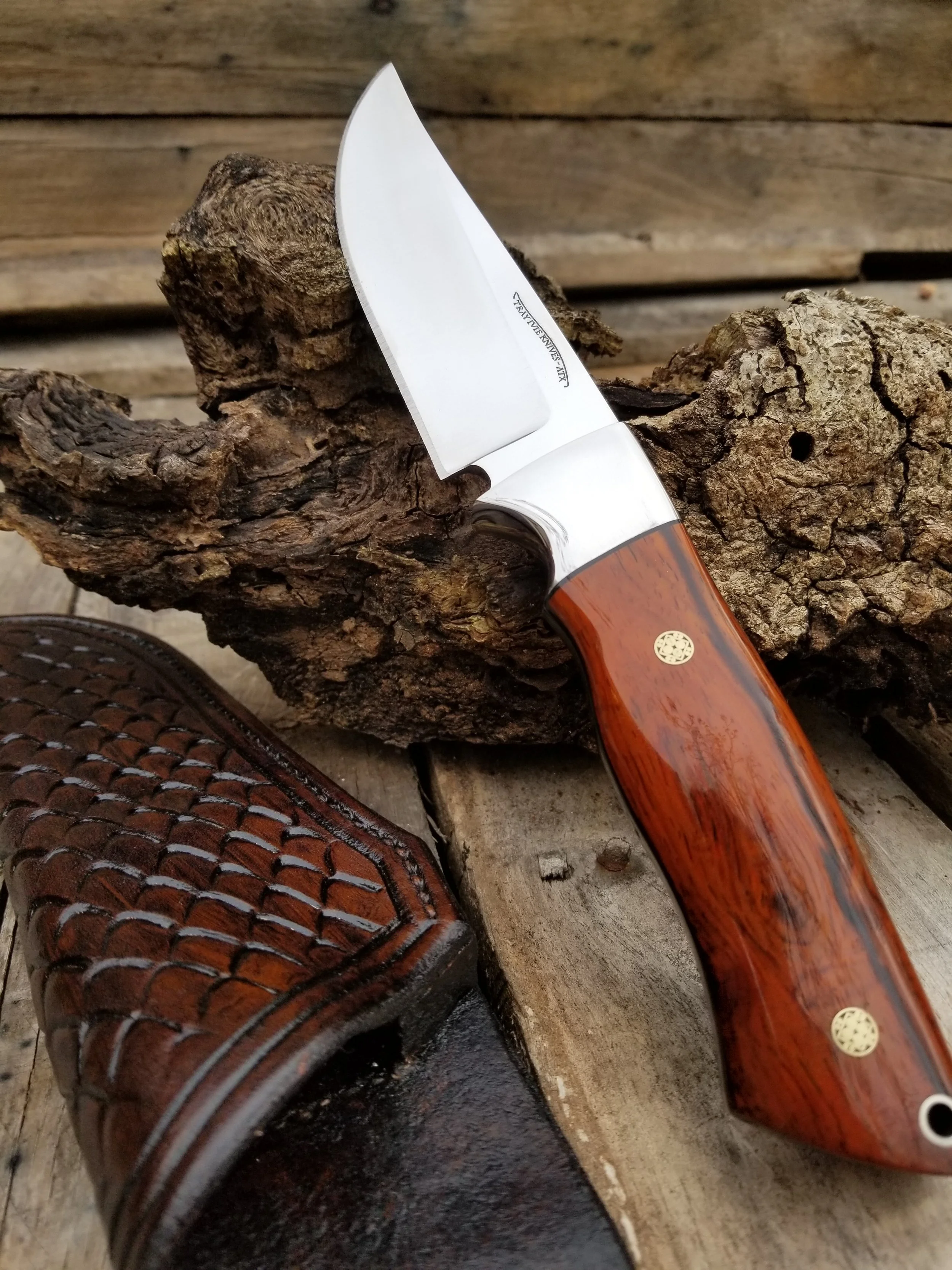 A hunting knife with a wooden handle resting on a piece of driftwood, with a leather sheath nearby on a wooden surface.