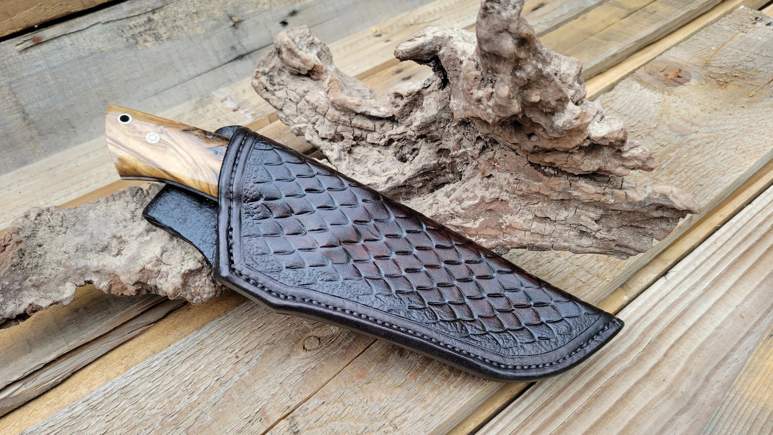 A fixed-blade knife with a wooden handle partially concealed in a black, textured, leather sheath, resting on logs with a piece of bark in the background.