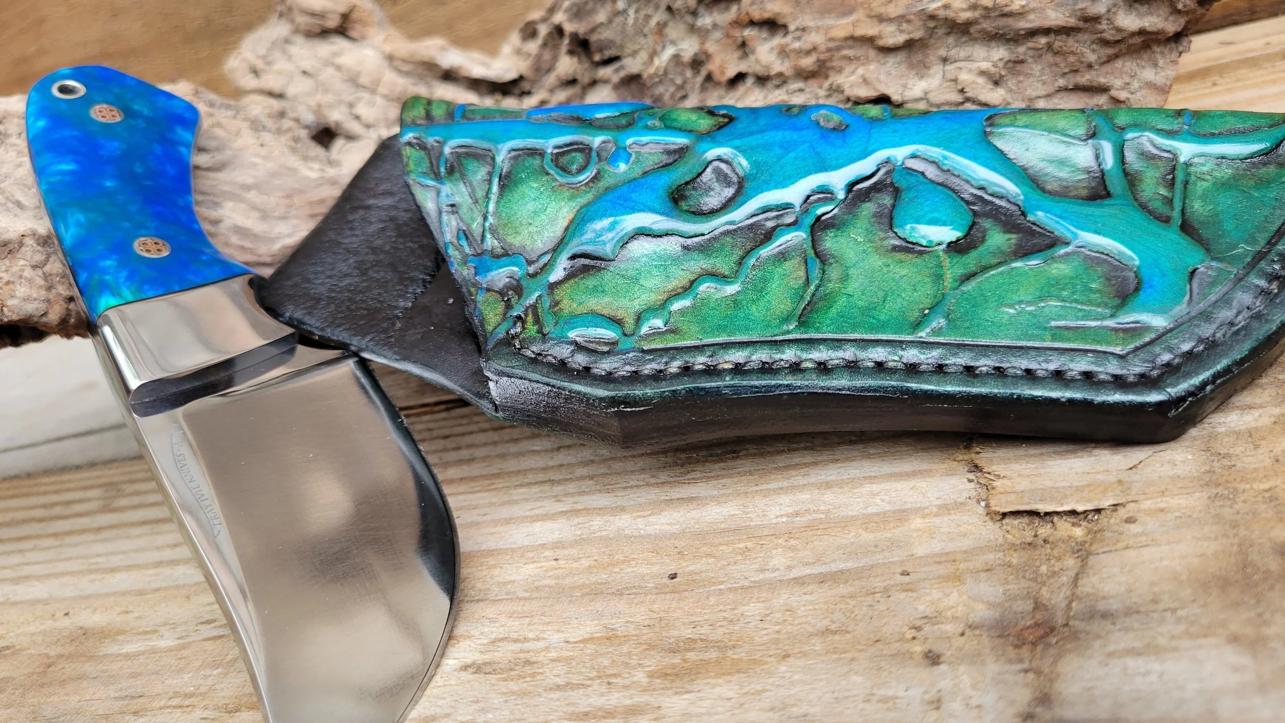 A folding pocket knife with a blue handle and stainless steel blade rests on a wooden surface. The handle is decorated with a mosaic inlay with green, blue, and black colors, and the knife is partly inside a black leather sheath.