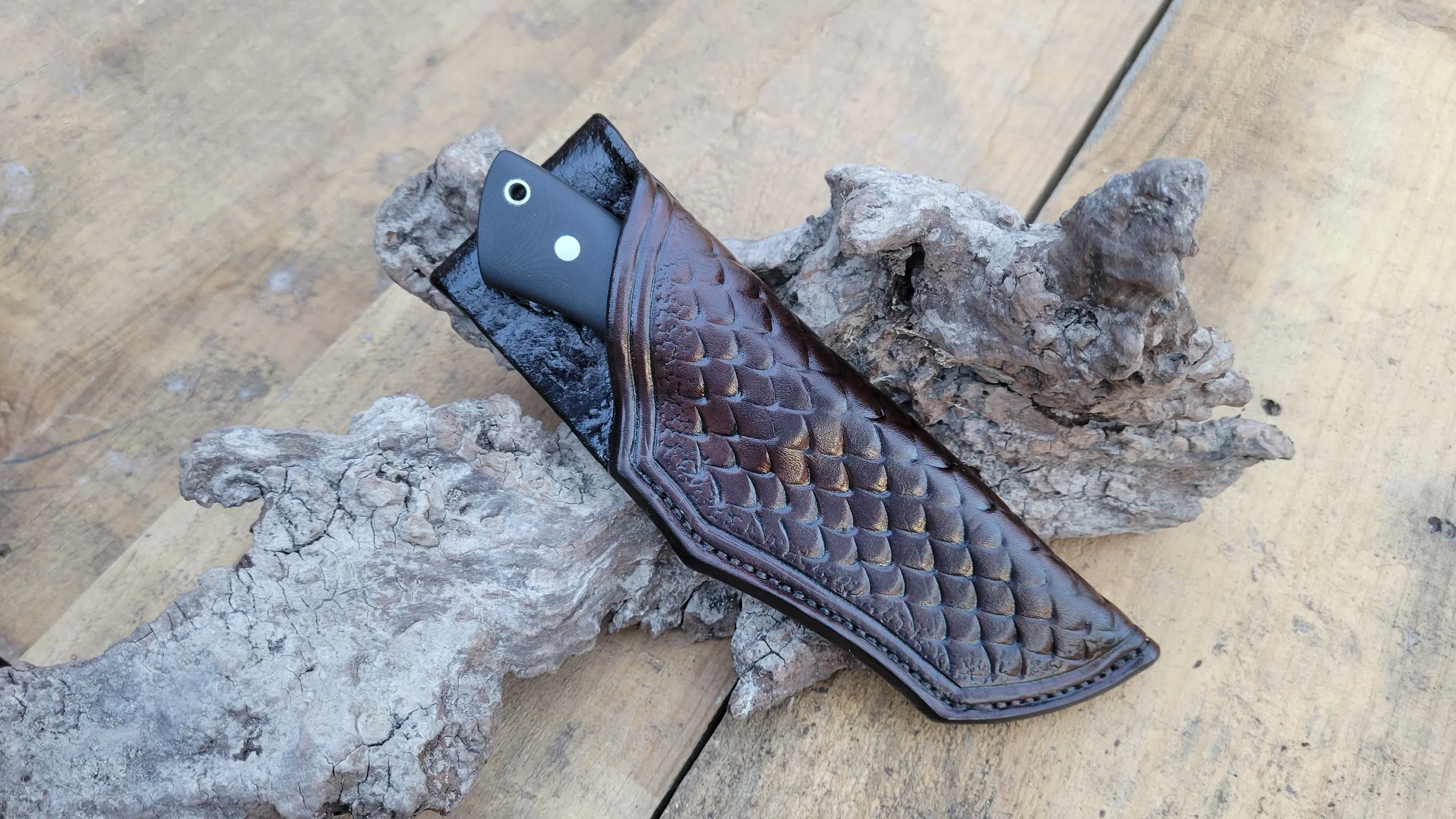 Close-up of a black-handled pocket knife in a leather sheath designed to resemble snakeskin, resting on a piece of textured, weathered wood.