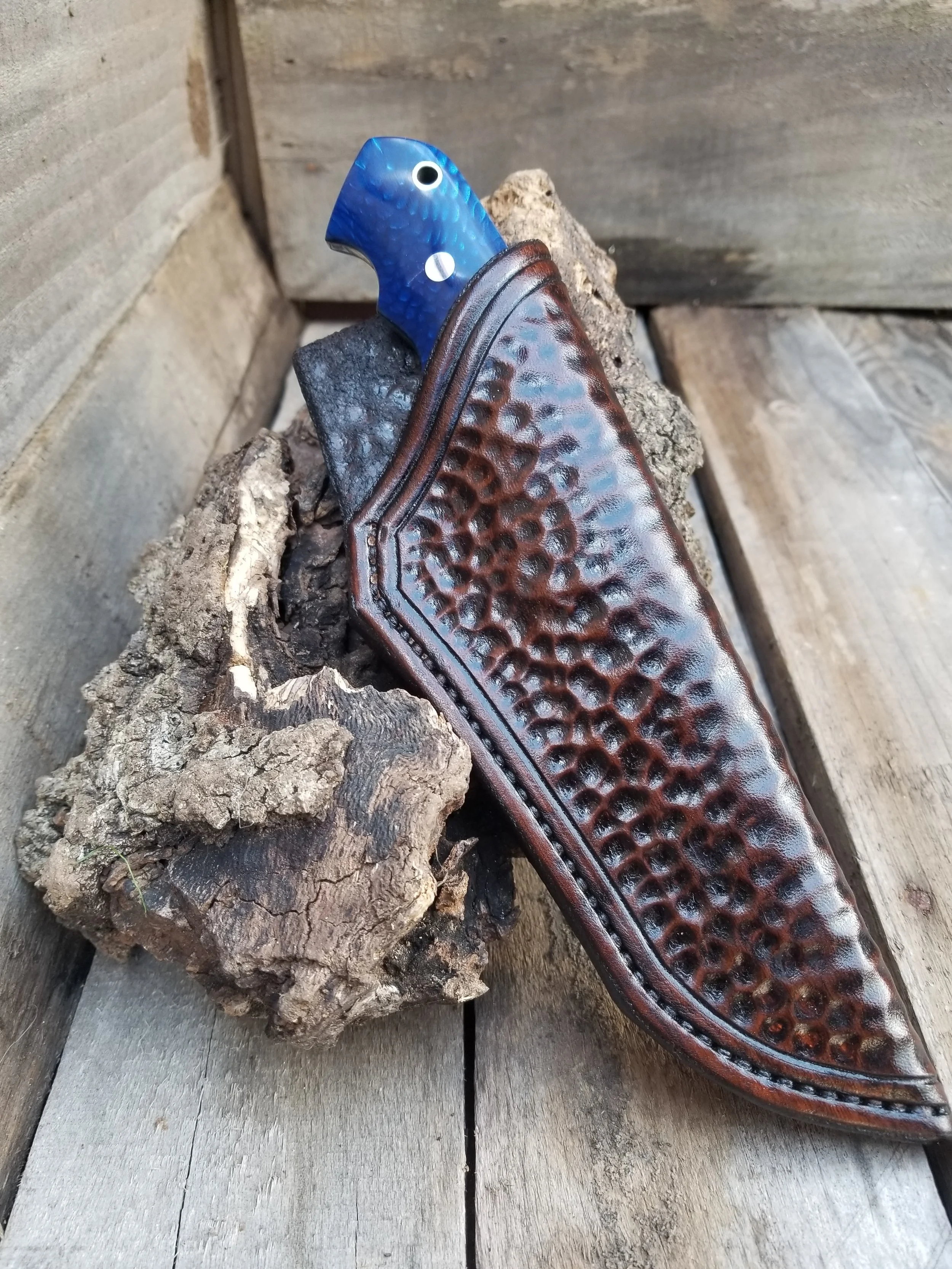 A knife with a blue fish-shaped handle in a brown leather sheath resting on a piece of wood on a rustic wooden surface.
