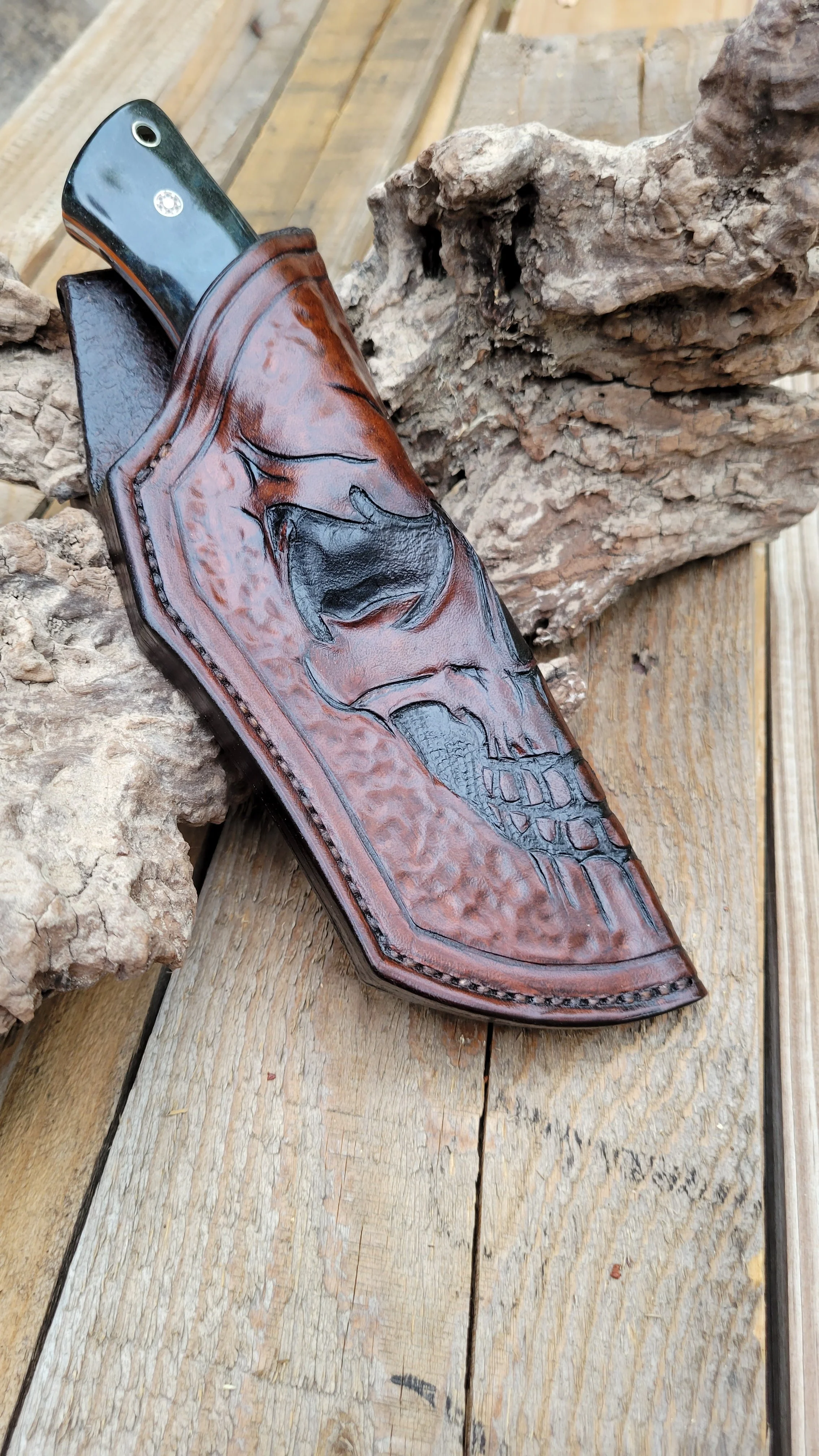 A knife with a handle attached to a leather sheath engraved with a skull design, placed on a wooden surface with pieces of wood around.