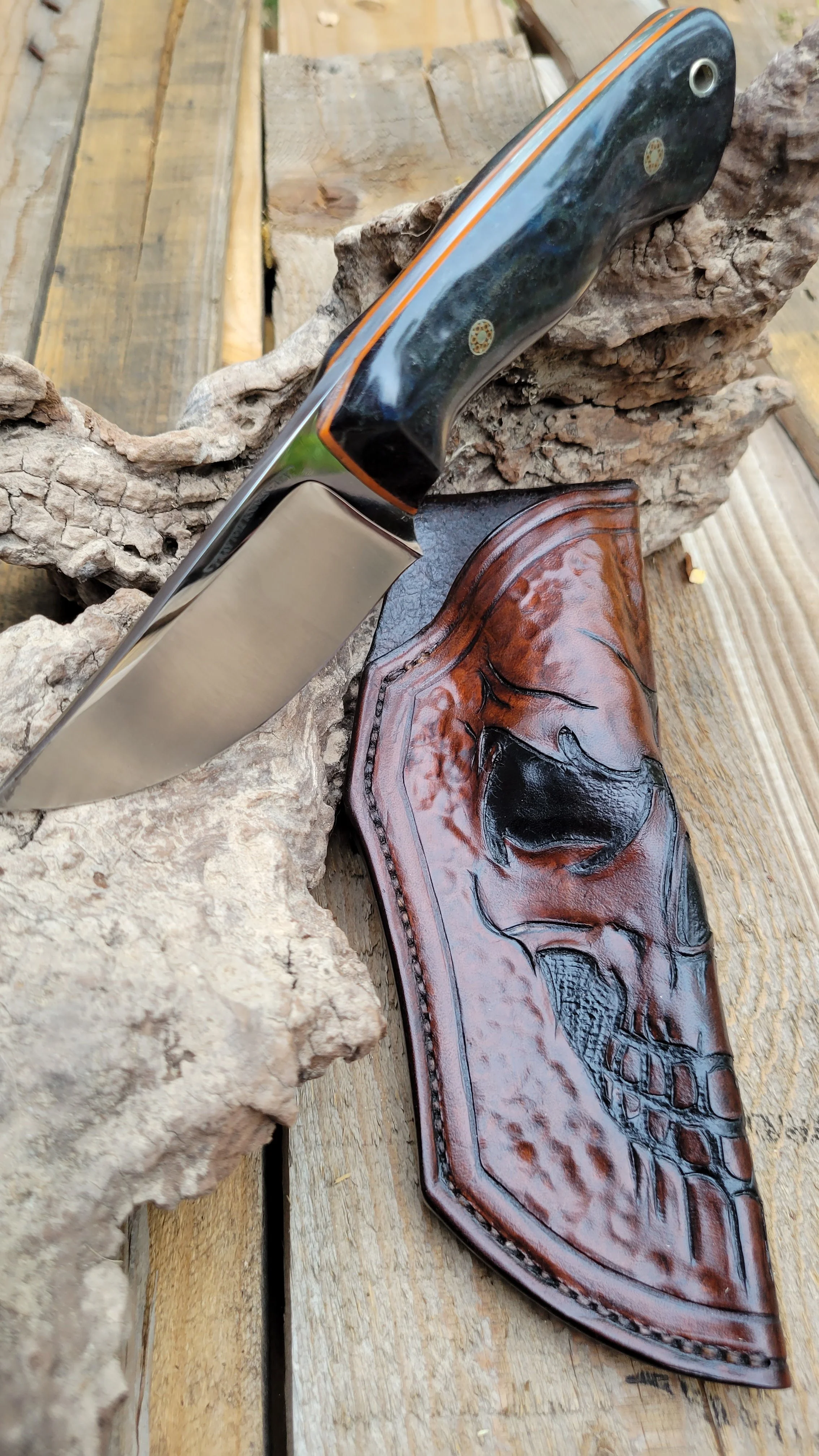 A hunting knife with a black handle and a shiny blade, resting on a piece of rough bark on a wooden surface, alongside a detailed leather sheath with a skull design.