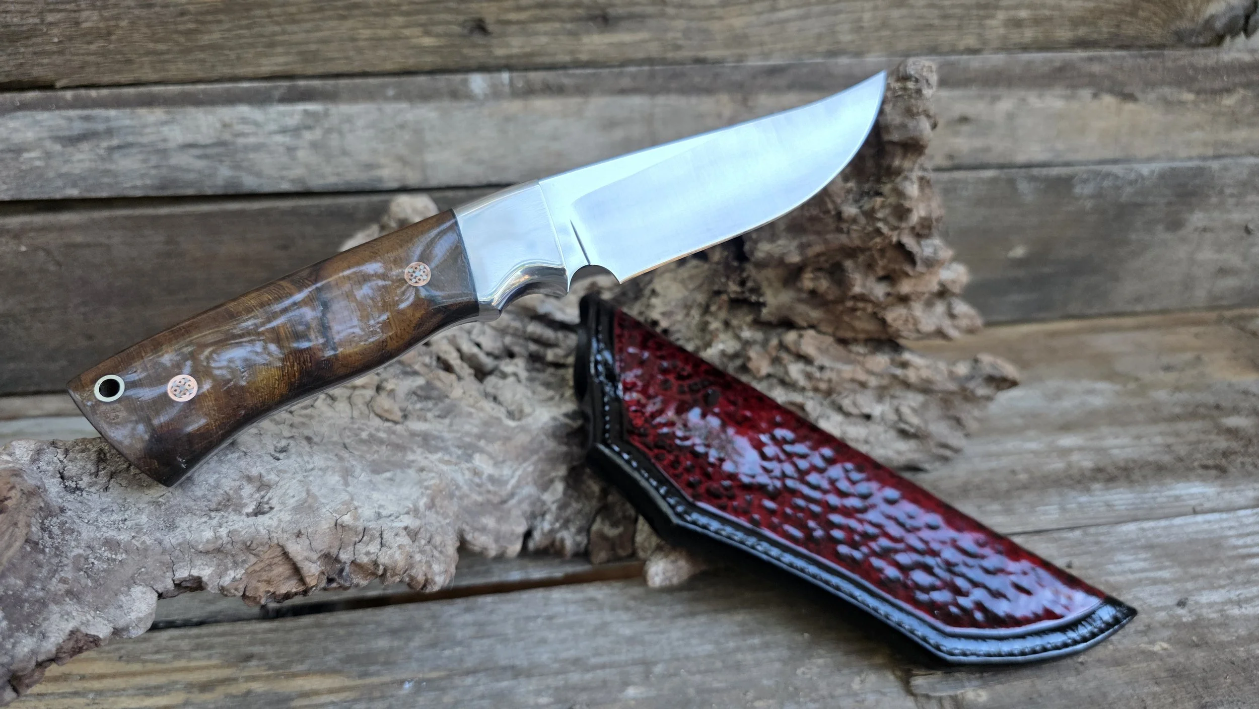 A folding pocket knife with a polished wooden handle and a stainless steel blade, resting on a piece of textured wood, with a red and black decorative case nearby.