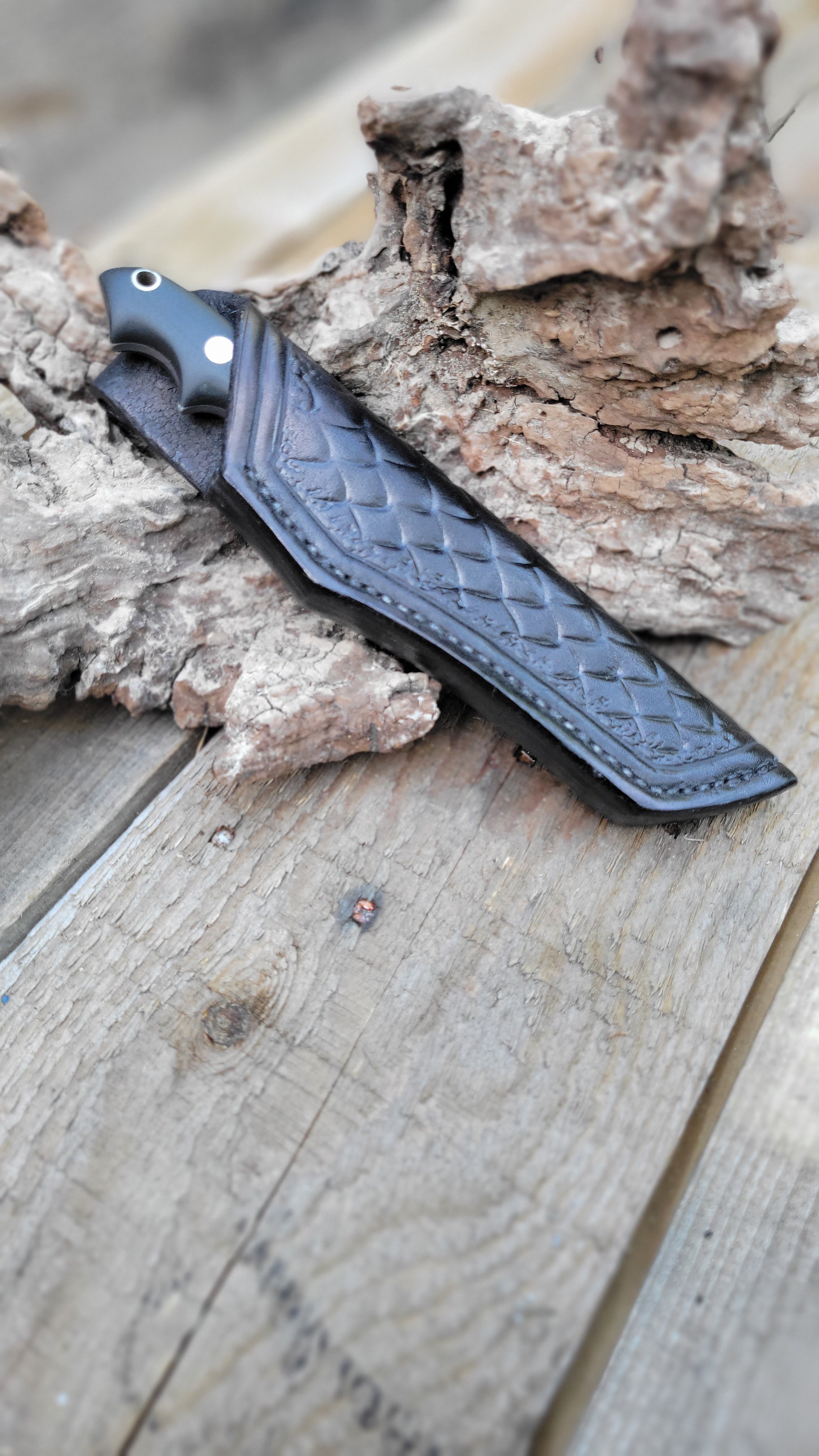 A black ceramic and metal knife with a textured handle lying on wooden planks next to pieces of rough, layered bark or wood.