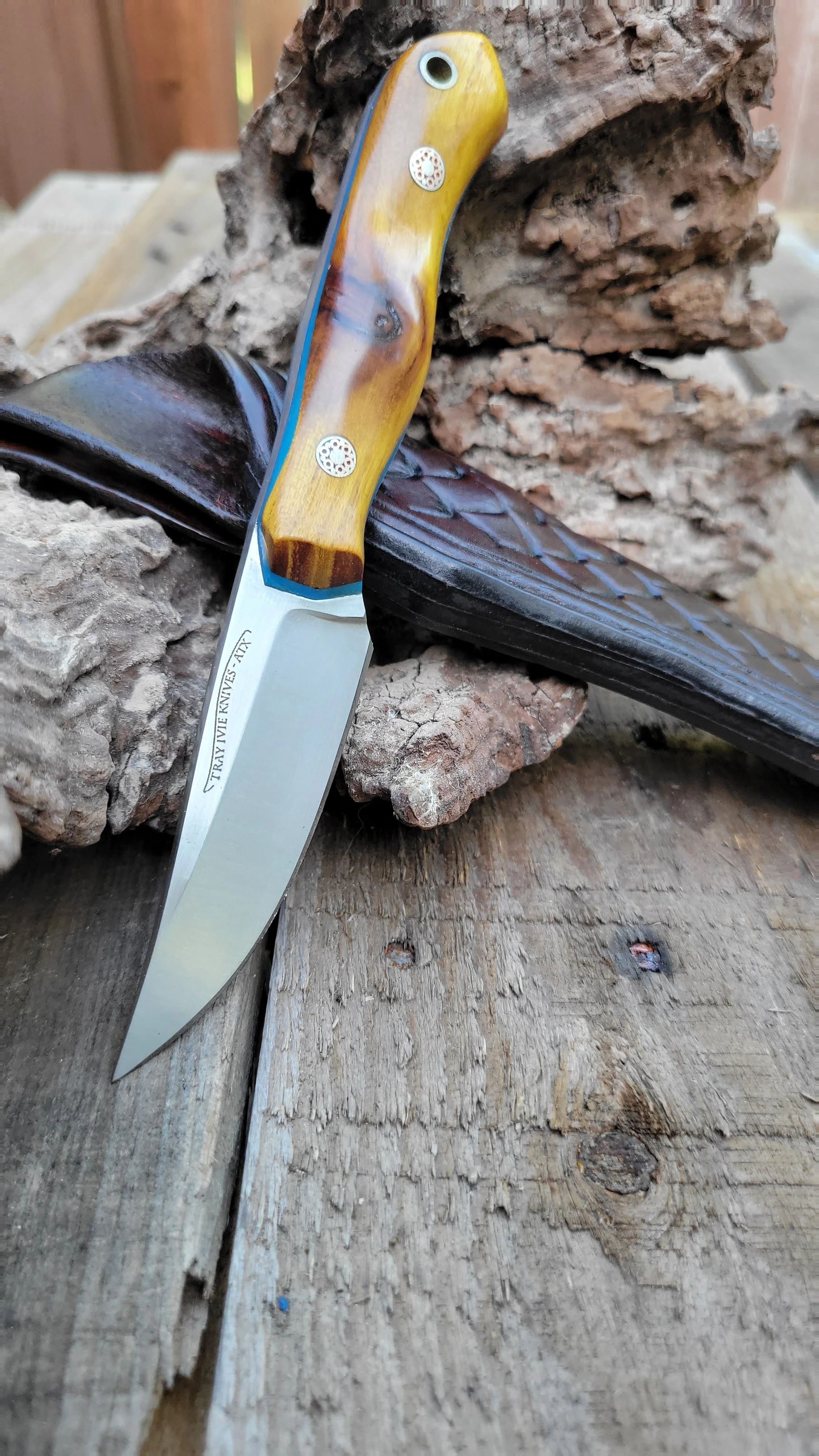 A fixed-blade knife with a yellow and brown wooden handle resting on a piece of bark and wood. The blade is inscribed with 'TRAY VIE KNIVES - ATX'.