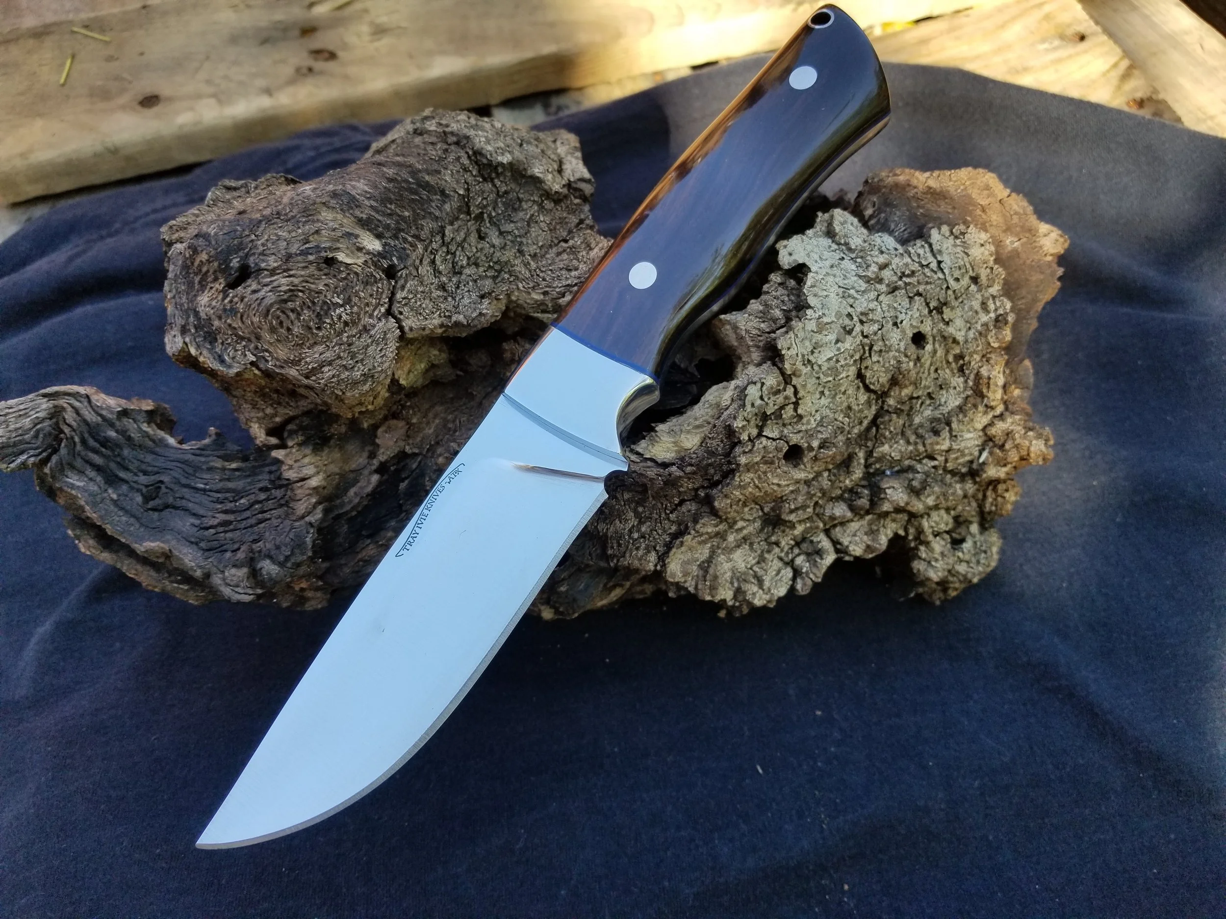 A kitchen knife with a dark wooden handle and a shiny metal blade is placed on a piece of rough, textured bark. The background includes a wooden surface and dark fabric.